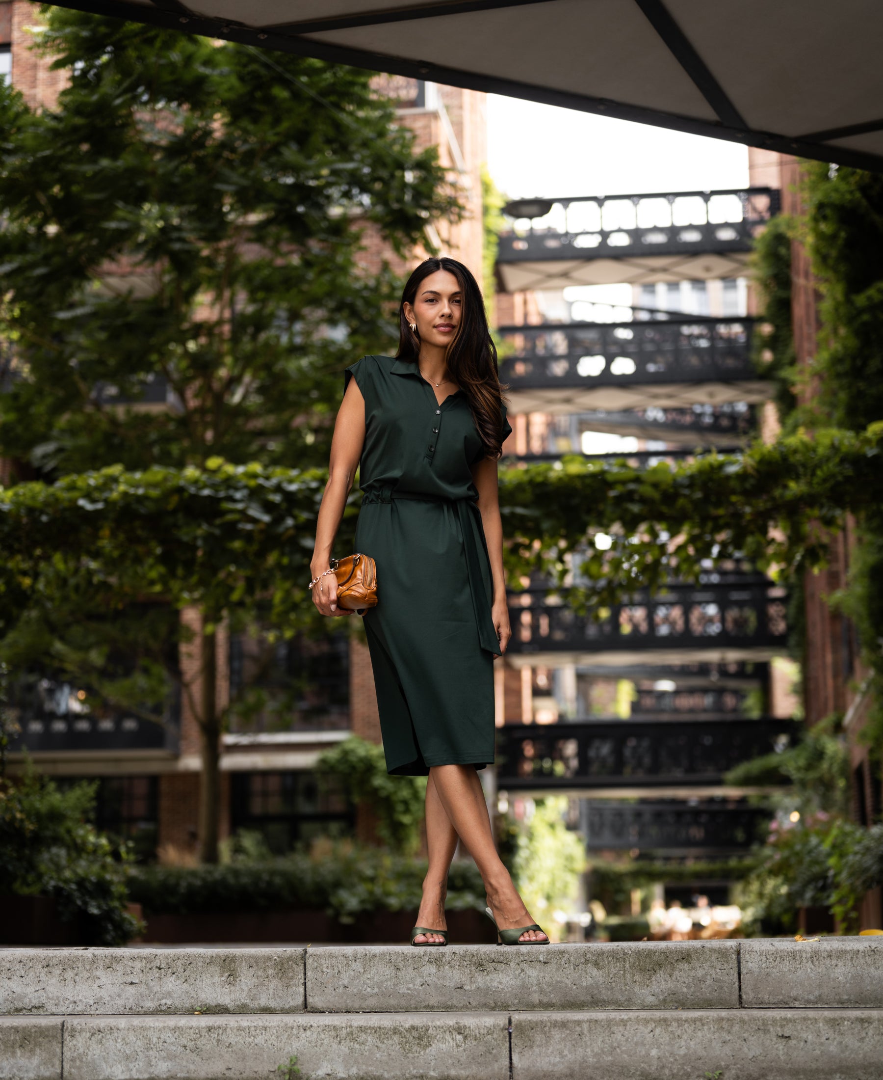 Woman wearing a dark green INES travel polo dress by PLEIN PUBLIQUE stands outdoors with city greenery in the background.