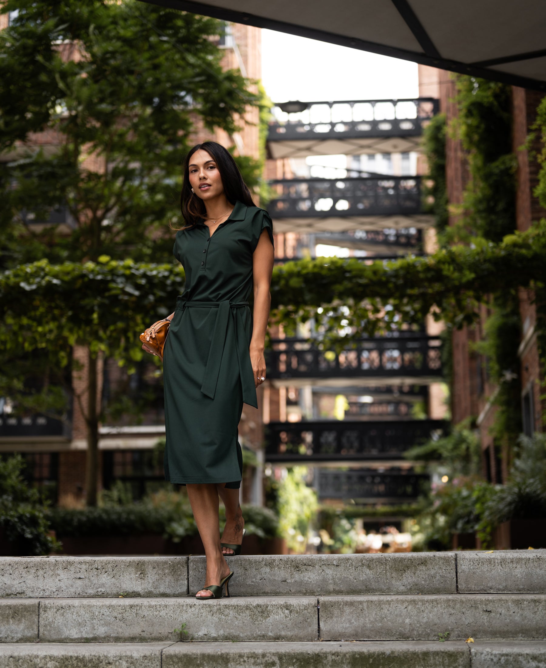 Woman wearing the PLEIN PUBLIQUE Travel polo dress INES in dark green walks outside with greenery and buildings behind.
