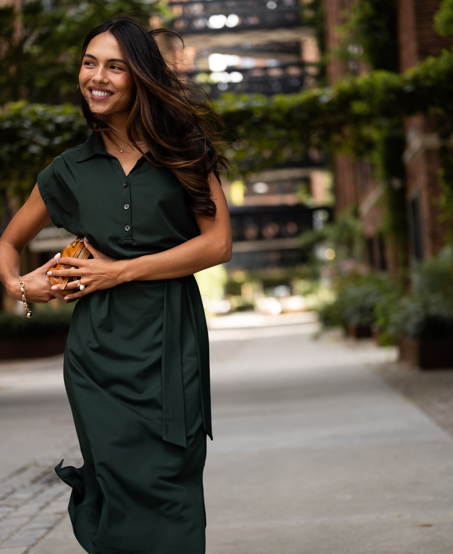 Woman wearing a dark green INES travel polo dress by PLEIN PUBLIQUE, shown walking outdoors on a sunny day.