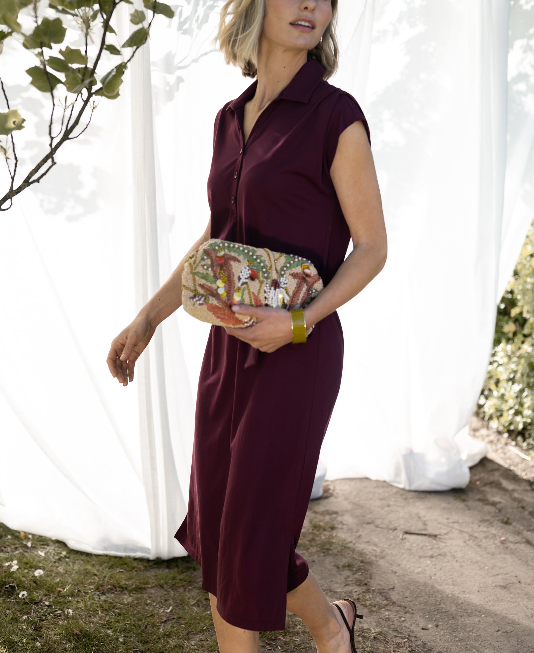 Woman wearing a wine-colored INES travel polo dress by PLEIN PUBLIQUE walks outdoors near greenery.