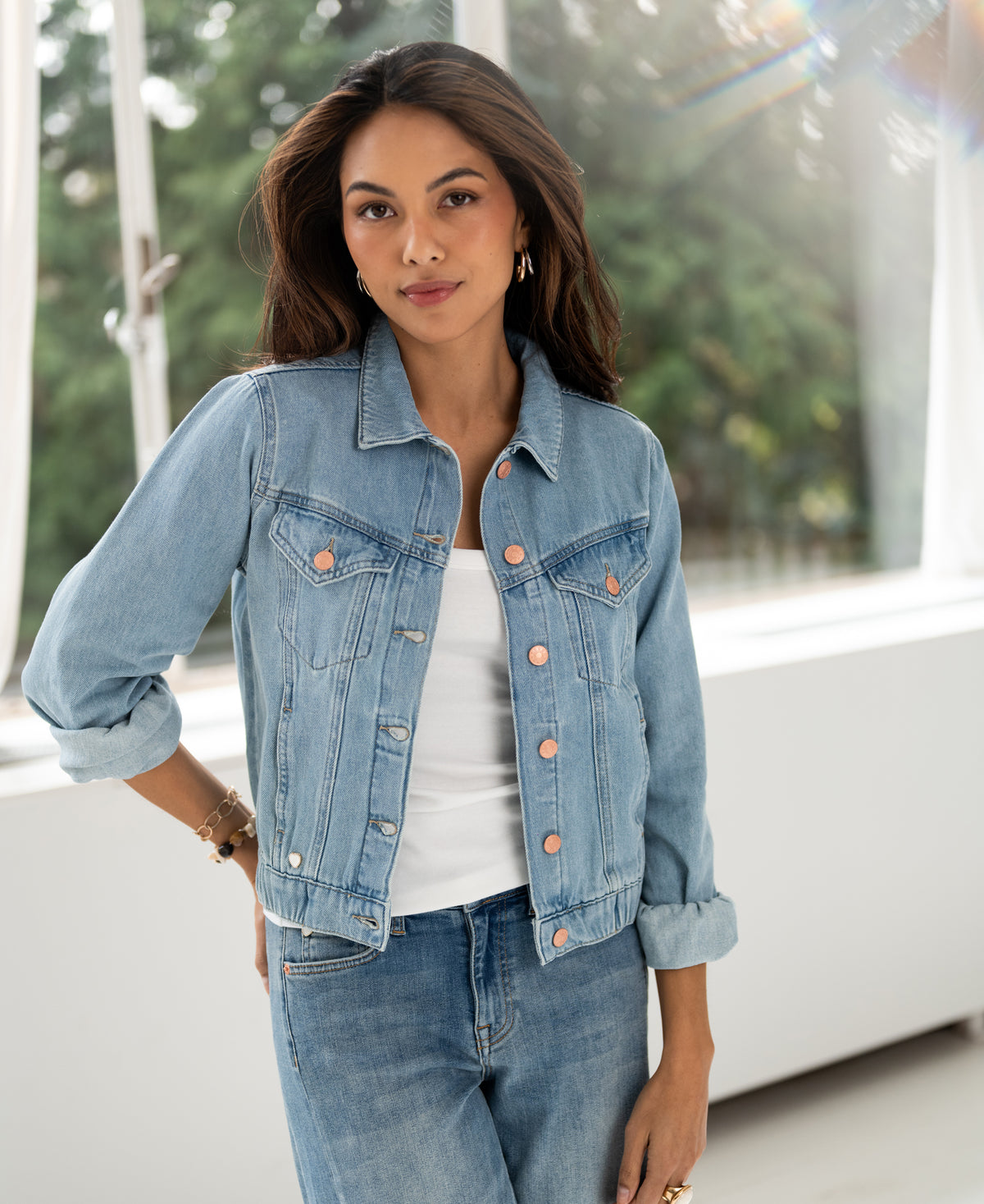 Woman wears bleached denim jacket LA JACKY indoors by a window with greenery visible outside.