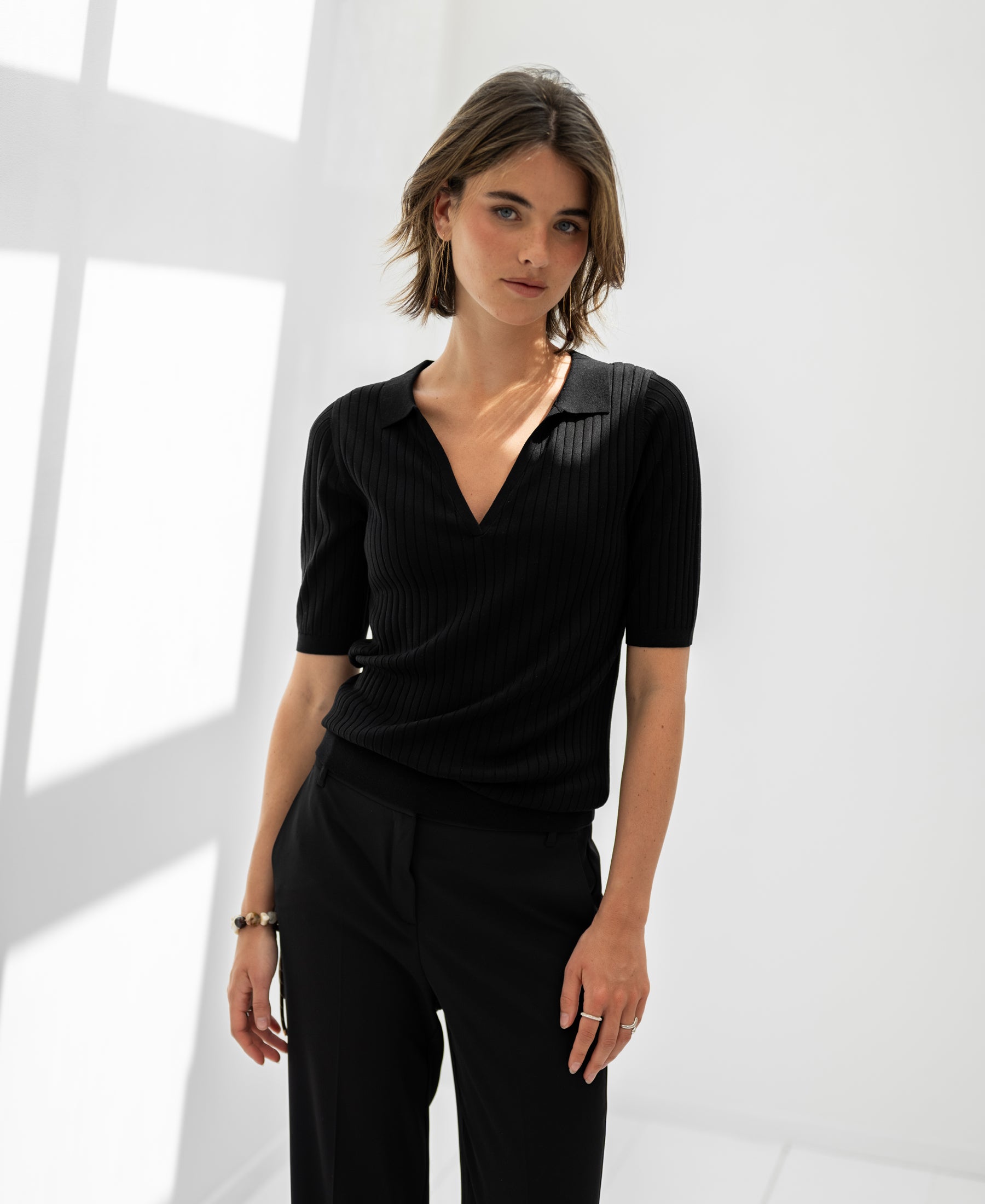 Woman wears black PLEIN PUBLIQUE Tencel-silk polo shirt in a bright room with white walls and window shadows.