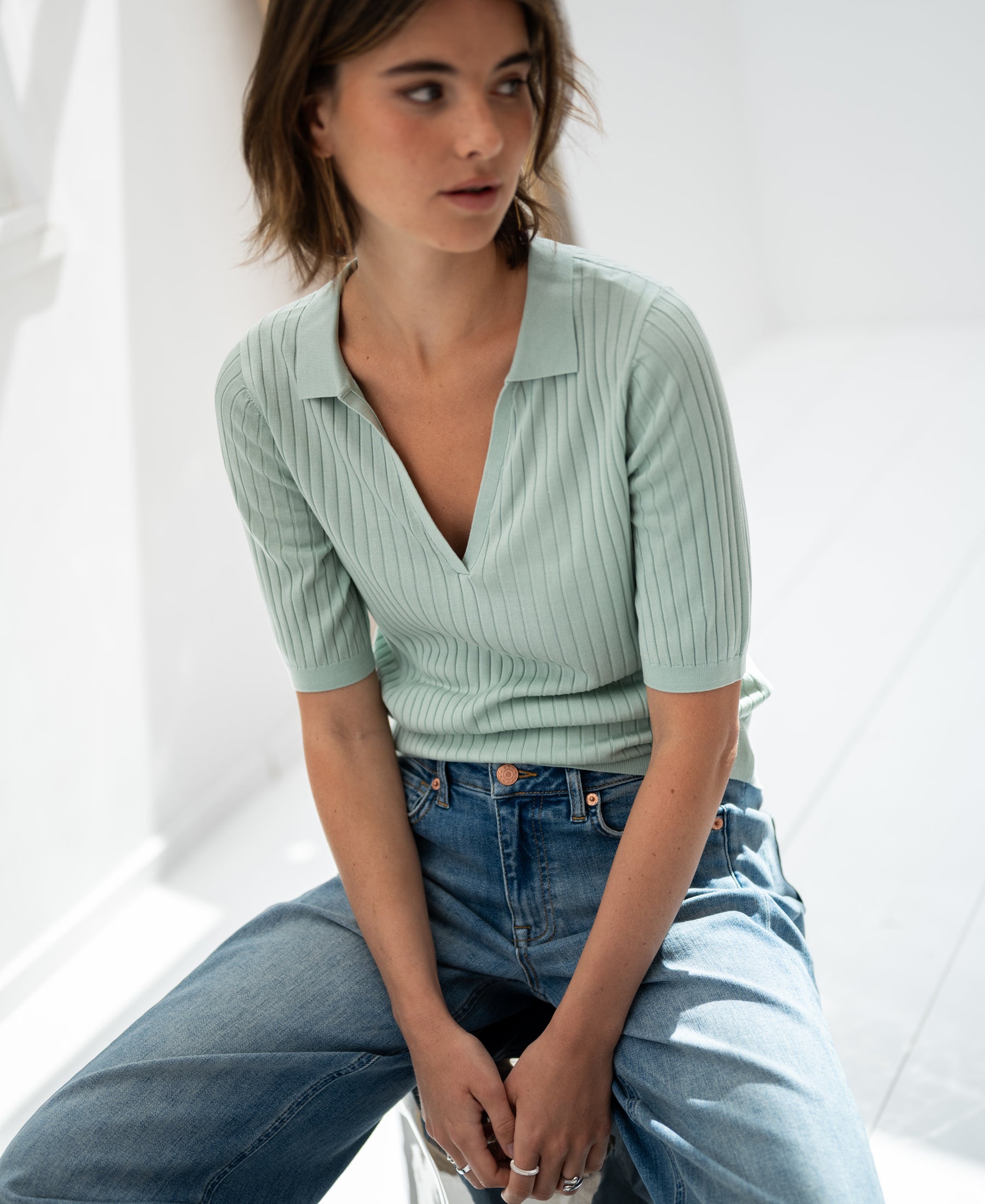 PLEIN PUBLIQUE’s Tencel-silk polo shirt in light green seen on a woman seated in a bright, minimal room.