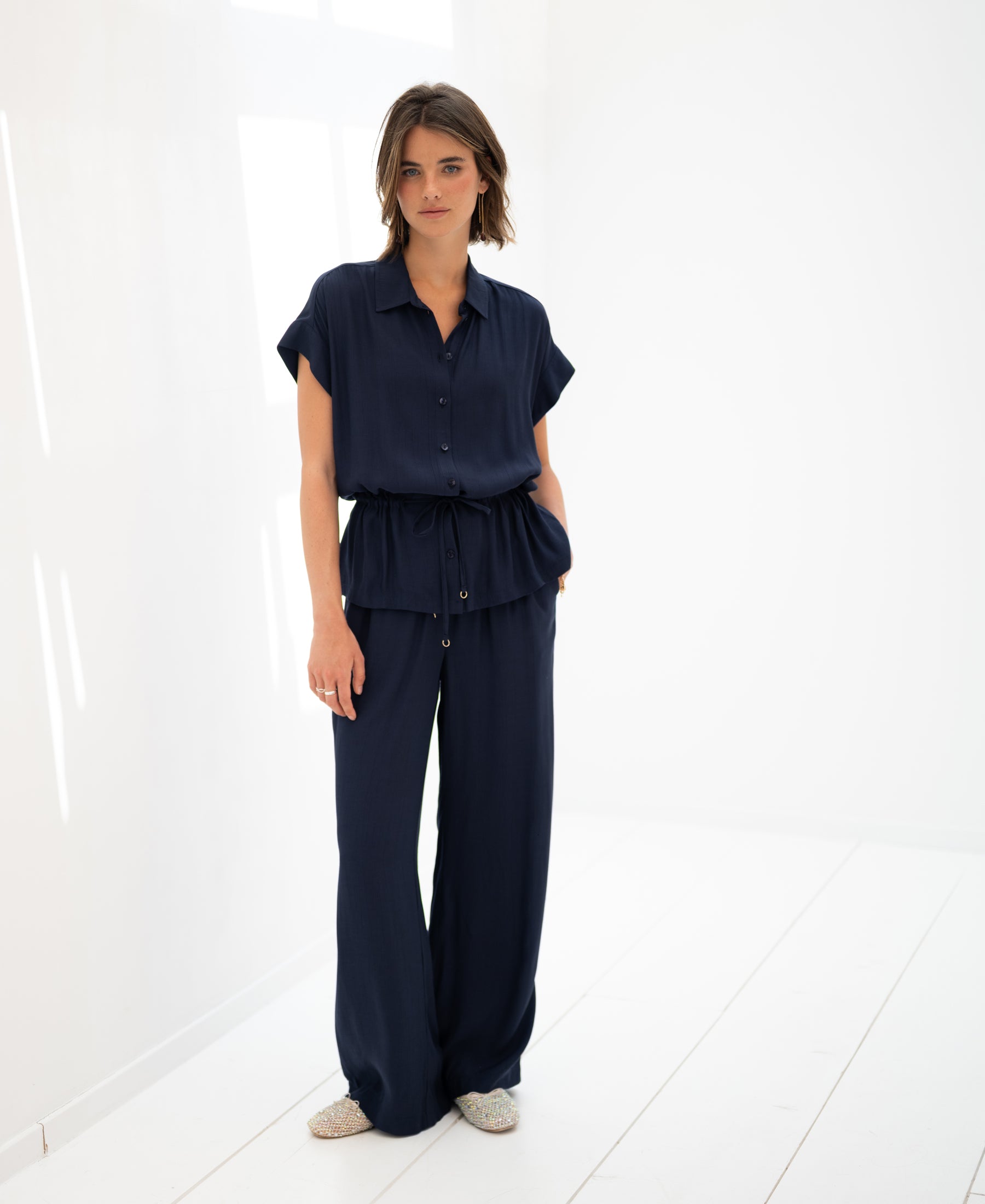 Dark blue LA LYNETTE trousers by OVERIG, shown from the front in a bright white minimalist room.
