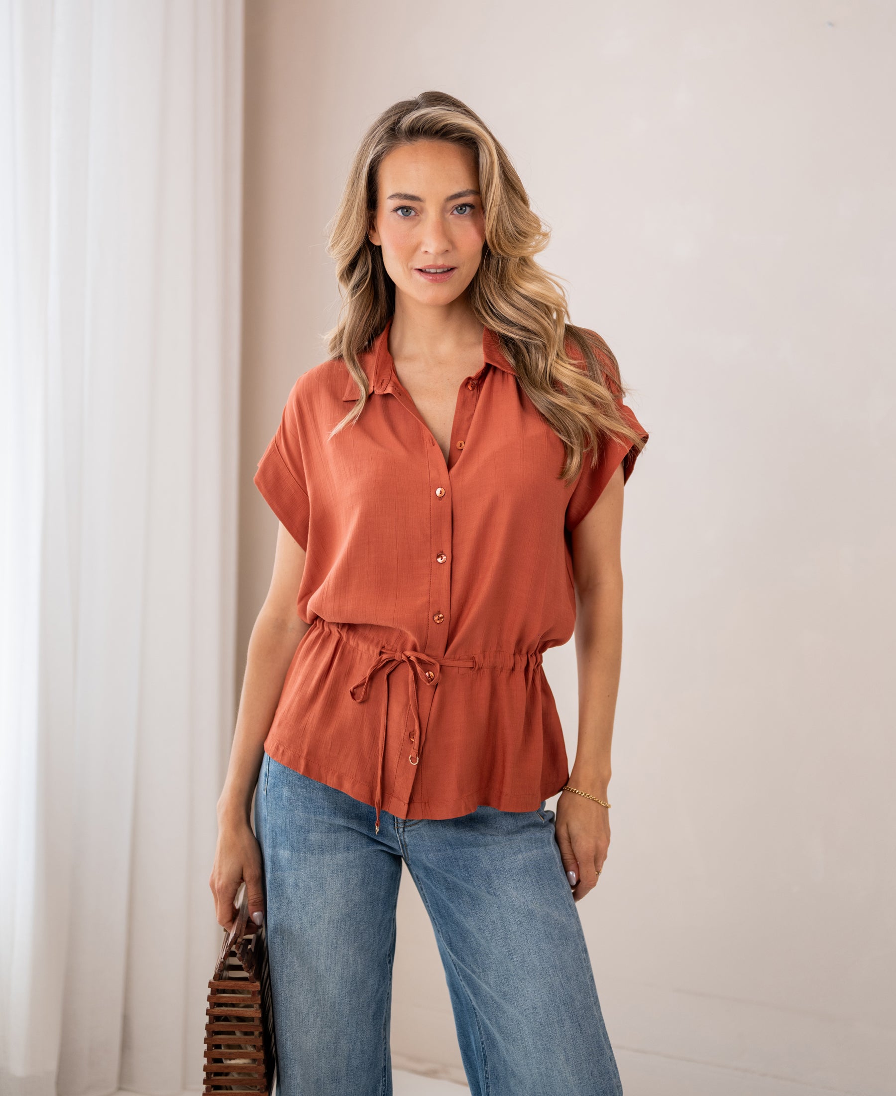 Woman wearing the LA JODY Terra blouse stands indoors with white curtains behind her.