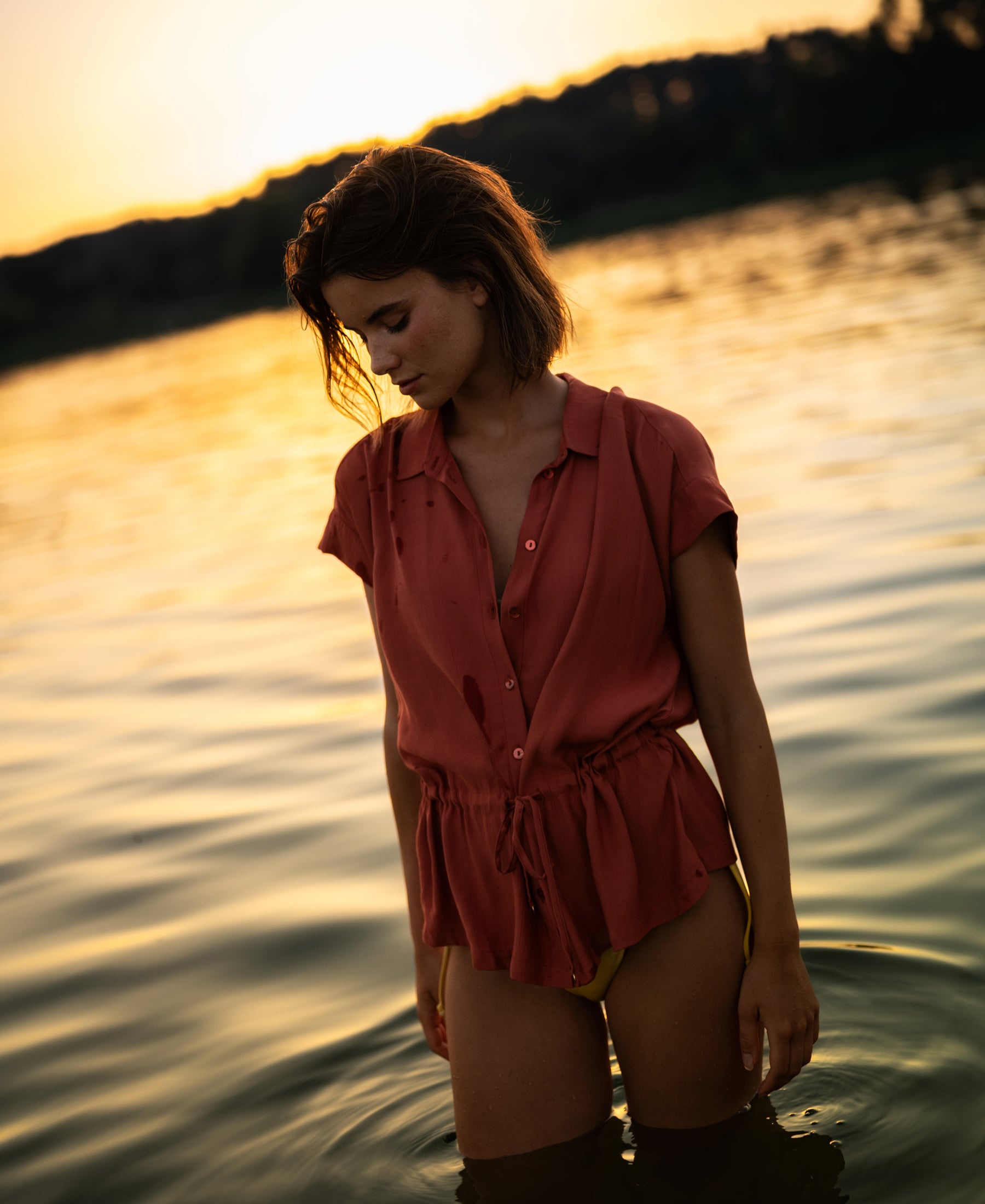Terra-colored LA JODY blouse by OVERIG displayed outdoors near water at sunset, trees in the background.