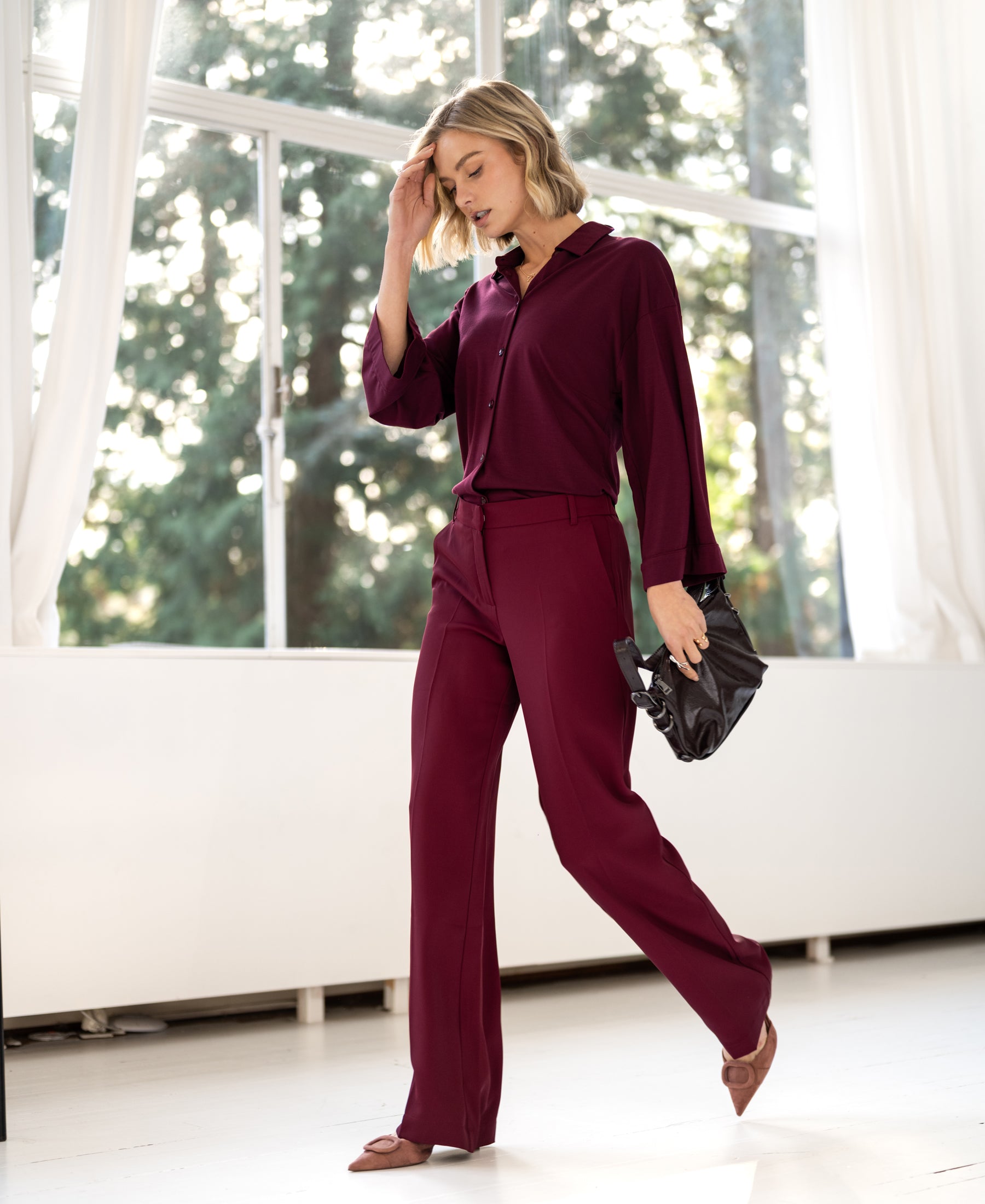 Woman in the flowy LA JOELLE trousers from PLEIN PUBLIQUE standing in sunlight near large windows.