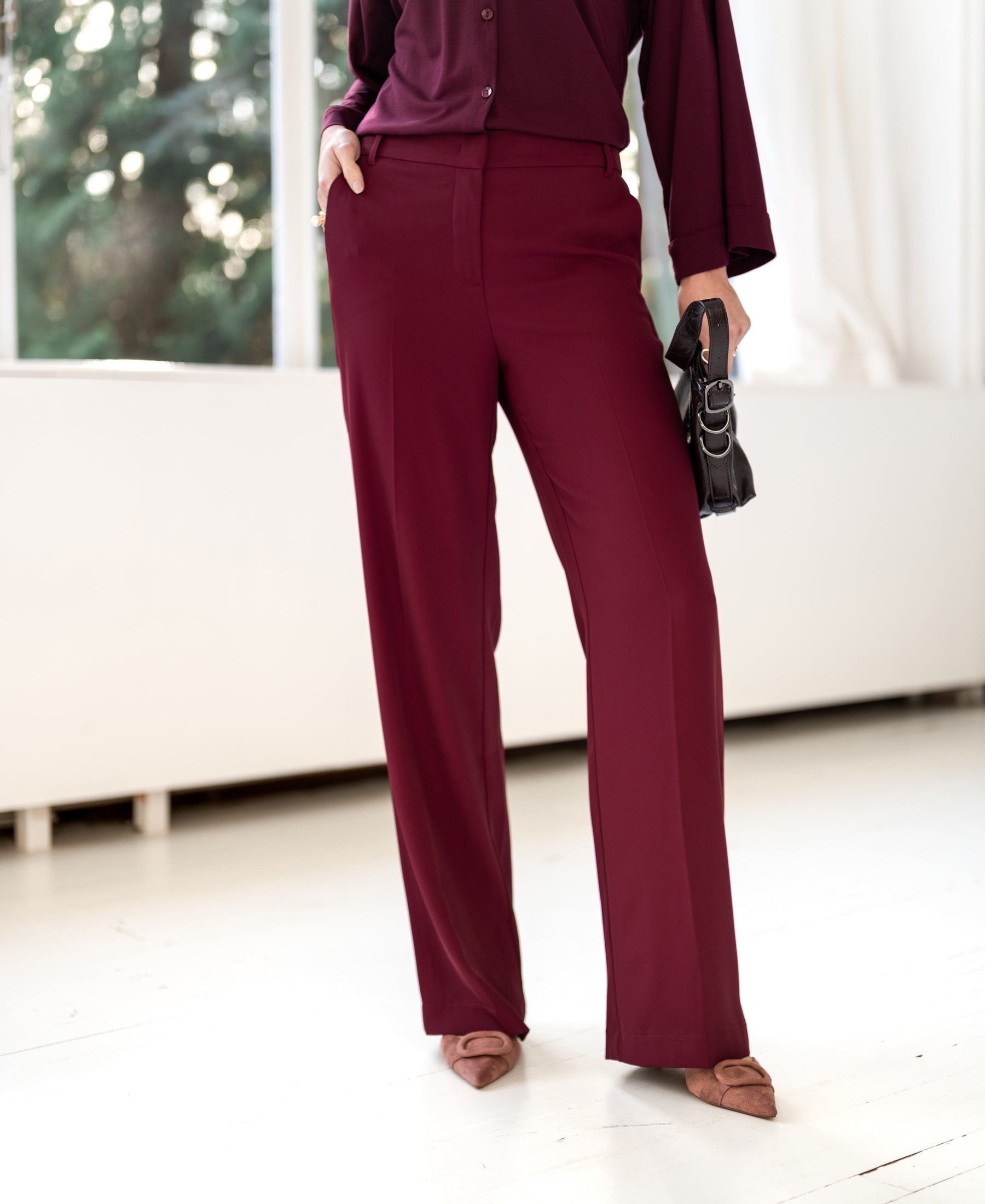 Vrouw draagt LA JOELLE Smooth Pants in Wine van PLEIN PUBLIQUE, poseert binnen bij een raam.