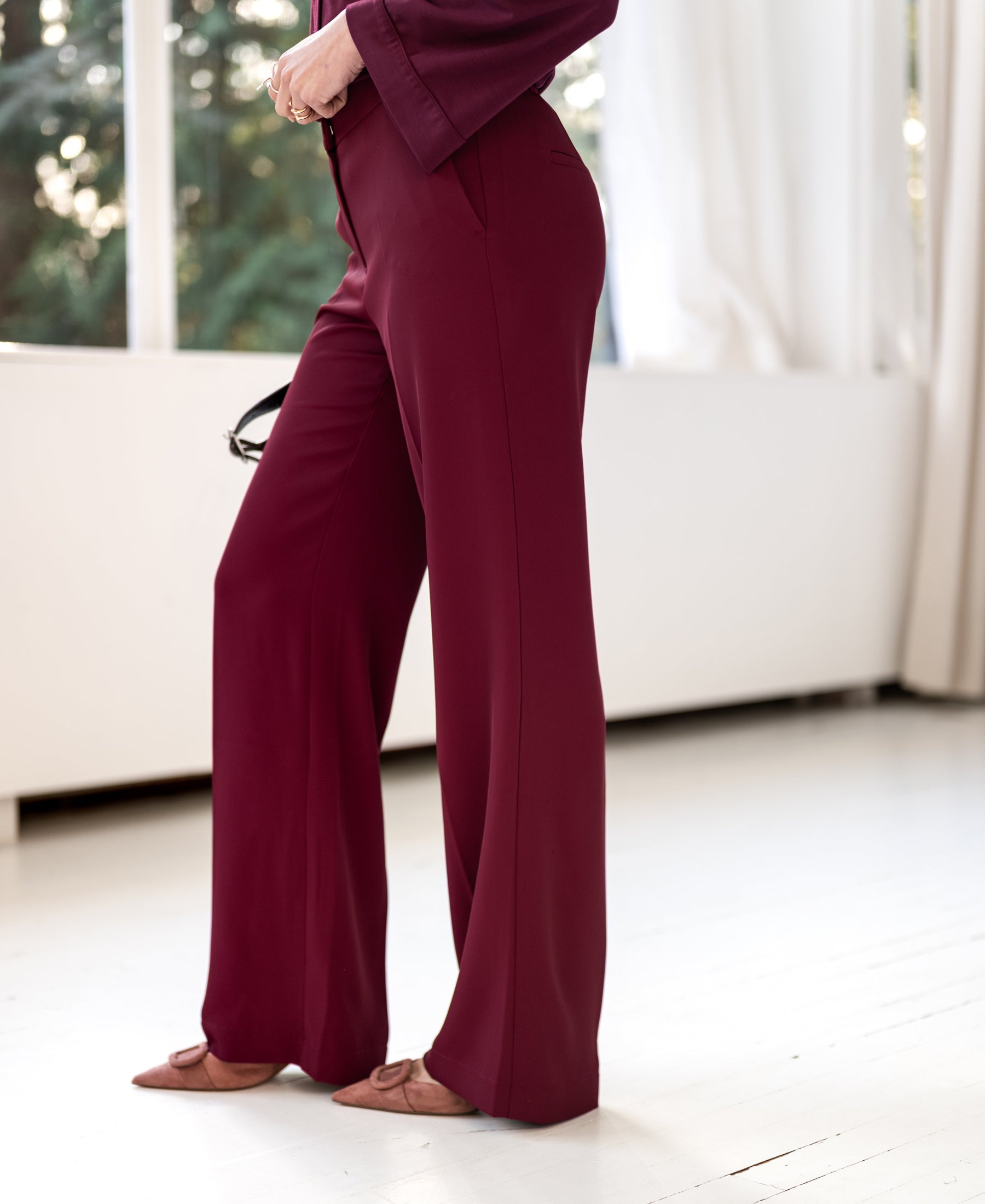 Woman wearing PLEIN PUBLIQUE LA JOELLE wine-colored supple pants near window with light curtains.