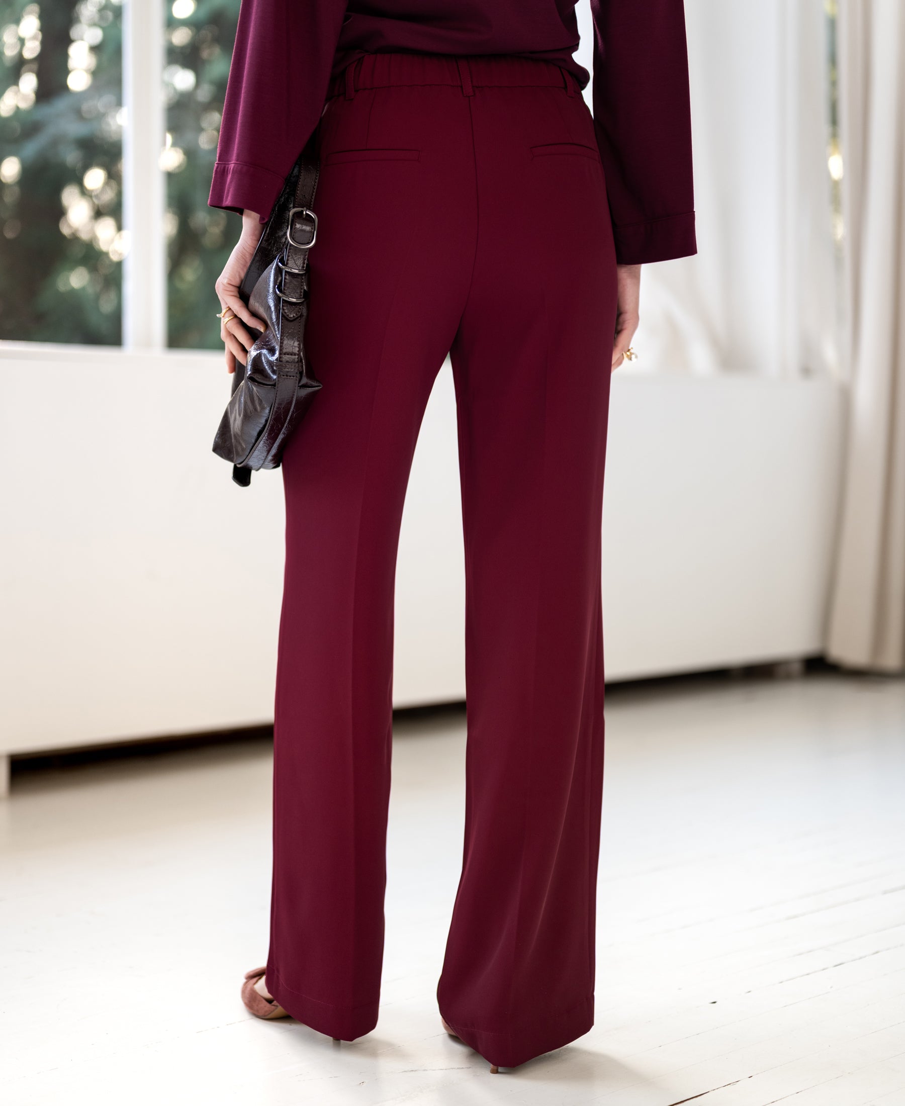 Woman wearing LA JOELLE Wine Smooth Pants from PLEIN PUBLIQUE, standing by large windows with white curtains.