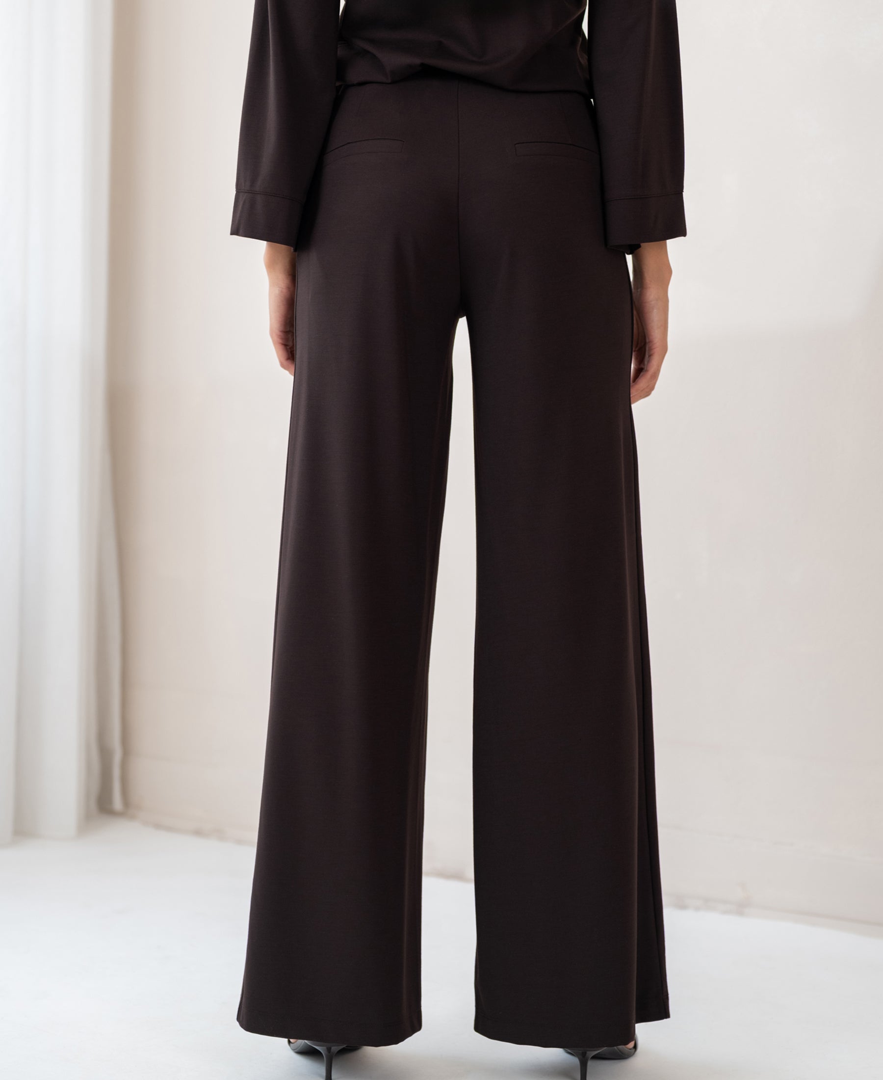 Back view of PLEIN PUBLIQUE LA JULIA Travel Pants in Pure Choco, worn by a person indoors.