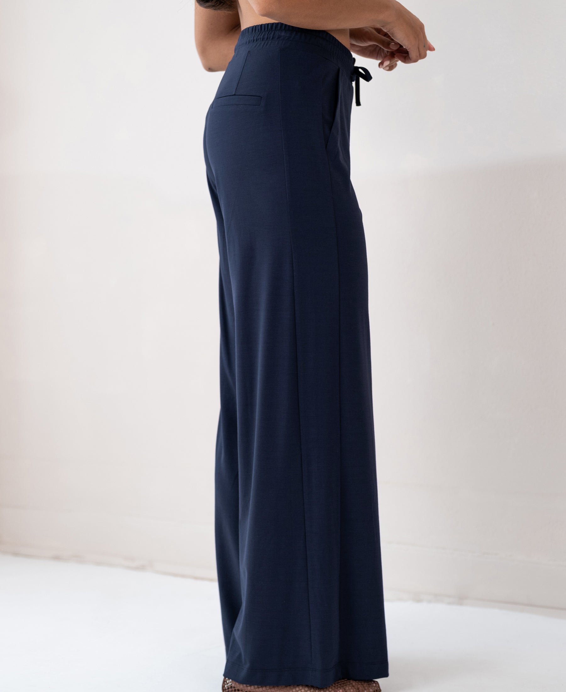Navy blue LA JULIA travel pants by PLEIN PUBLIQUE, shown from the side against a light wall.