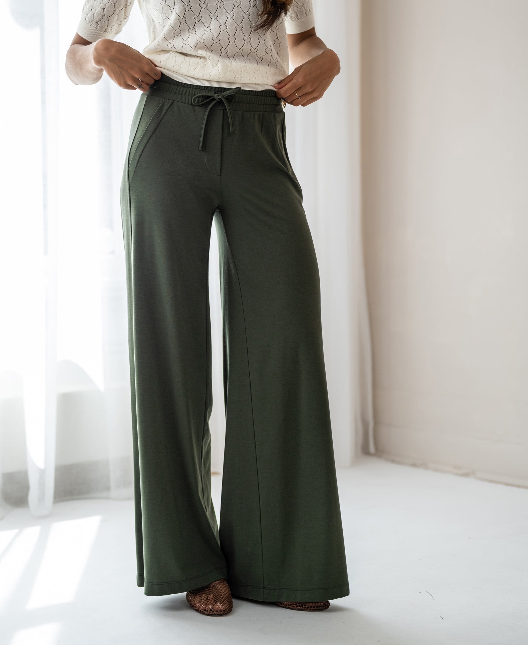 Olive green PLEIN PUBLIQUE Travel pants, style LA JULIA, shown on a woman standing in a bright room.
