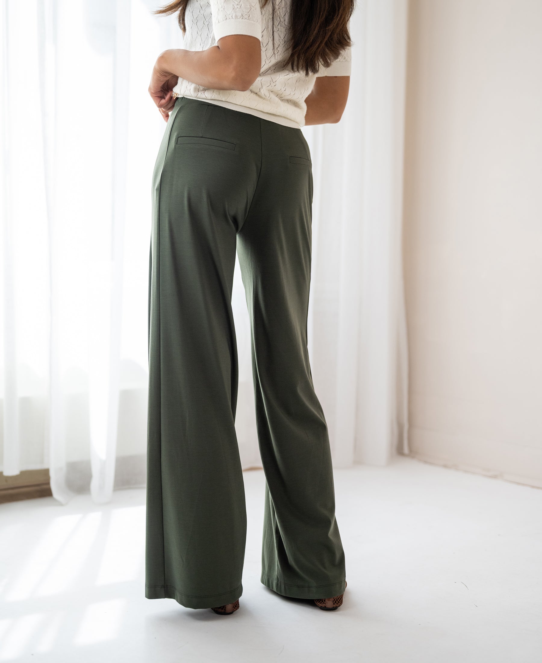 Dark olive travel pants by PLEIN PUBLIQUE displayed near a window with sheer curtains.