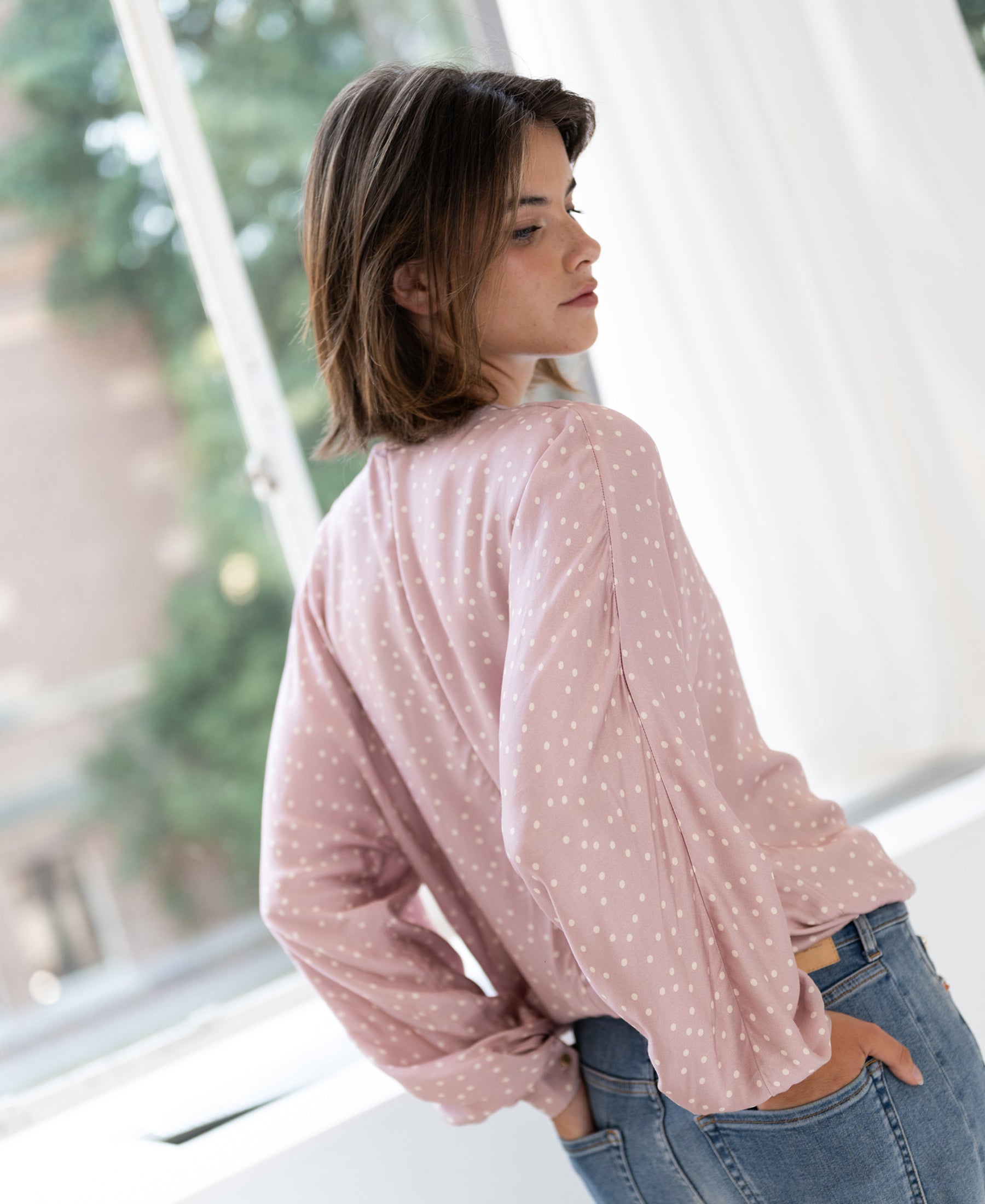 Woman wearing dusty pink and ivory polka dot wrap blouse stands by a window, looking back.