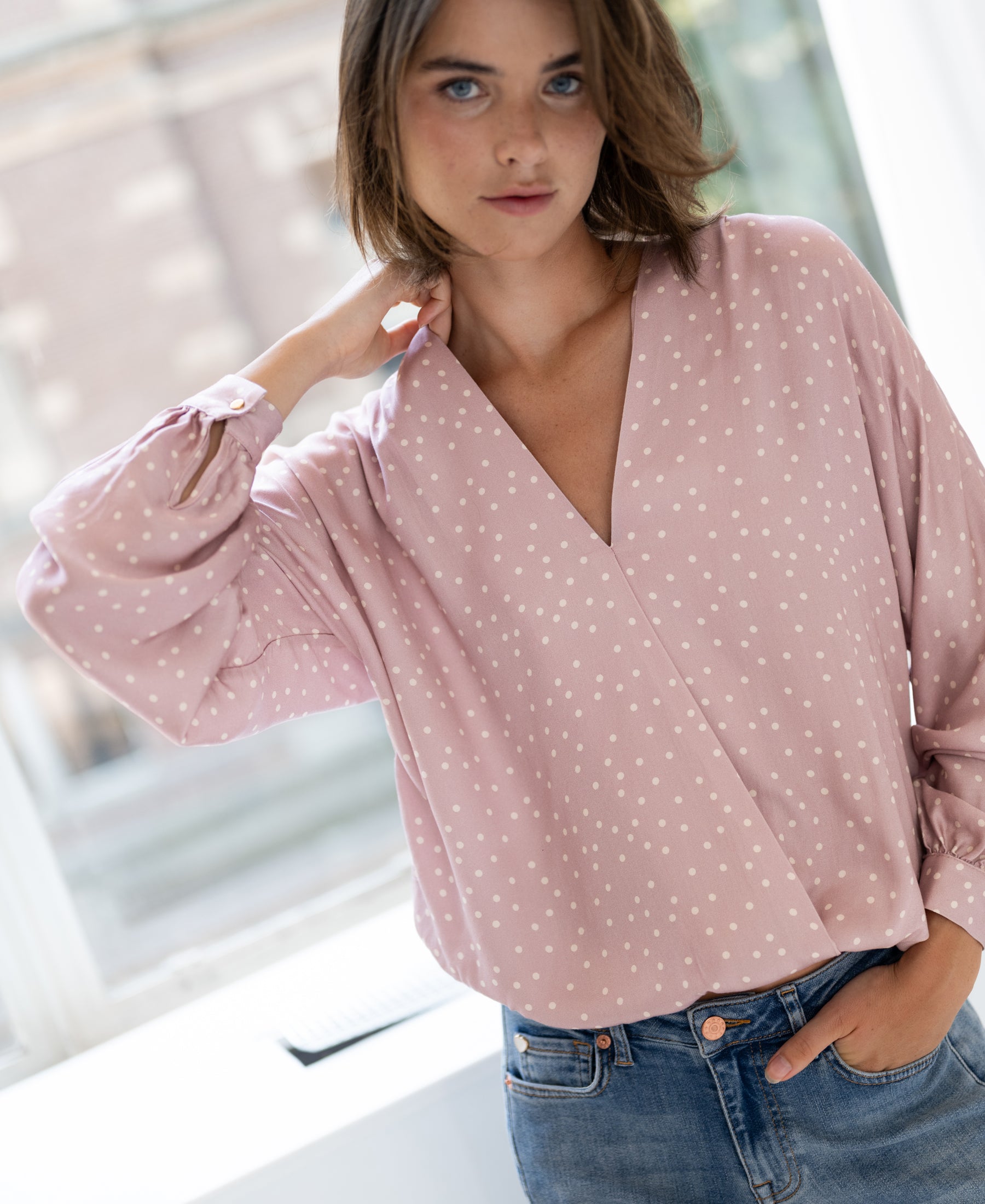 Woman wearing a Plein Publique LA LARA wrap blouse, pink with ivory polka dots, standing indoors with hand on neck.