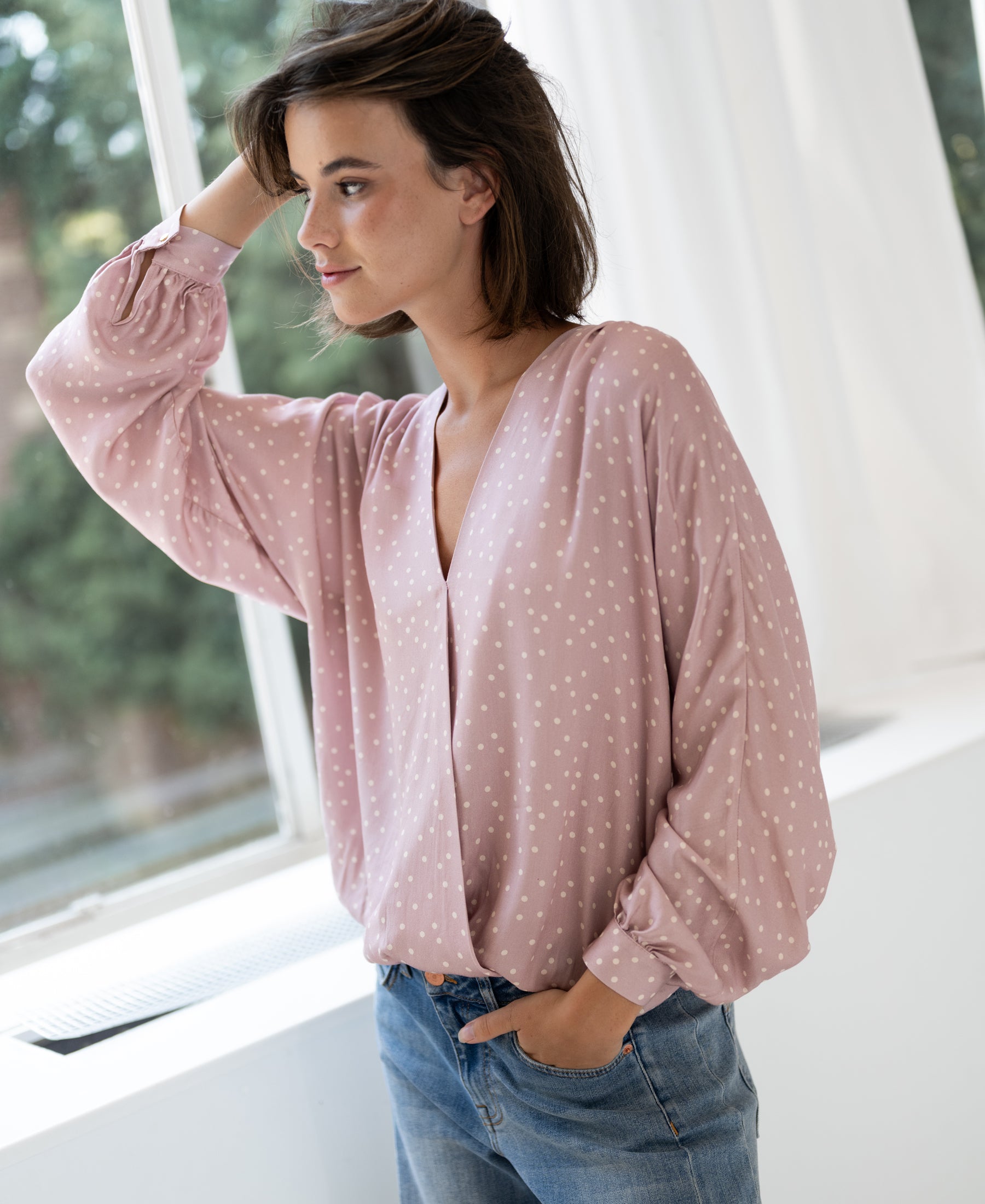 Dusty pink wrap blouse with dot pattern by PLEIN PUBLIQUE shown on model standing by a window, hand in pocket.