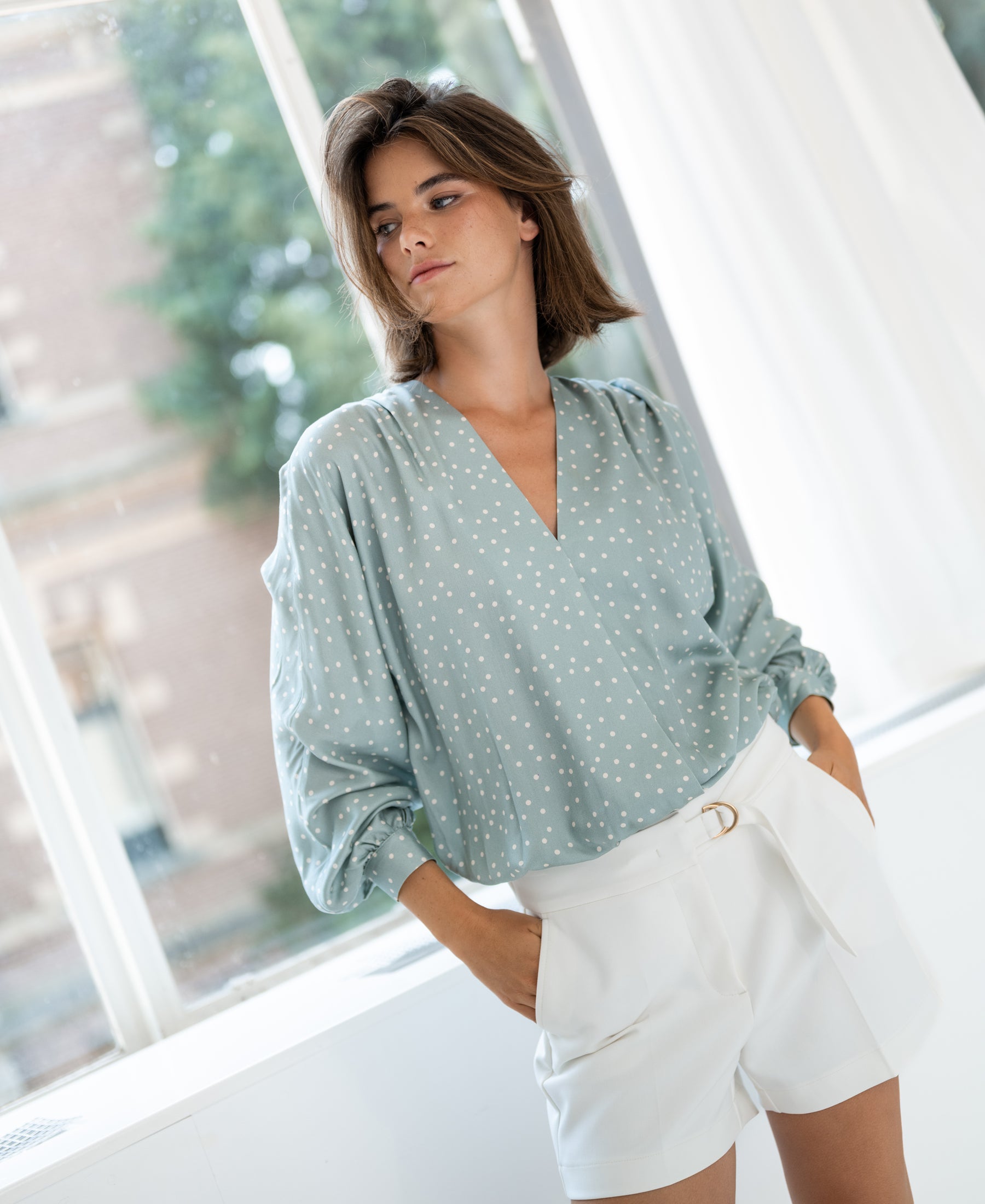 Woman wearing the PLEIN PUBLIQUE LA LARA Dot Mist Green - Ivory wrap blouse stands by a bright window.