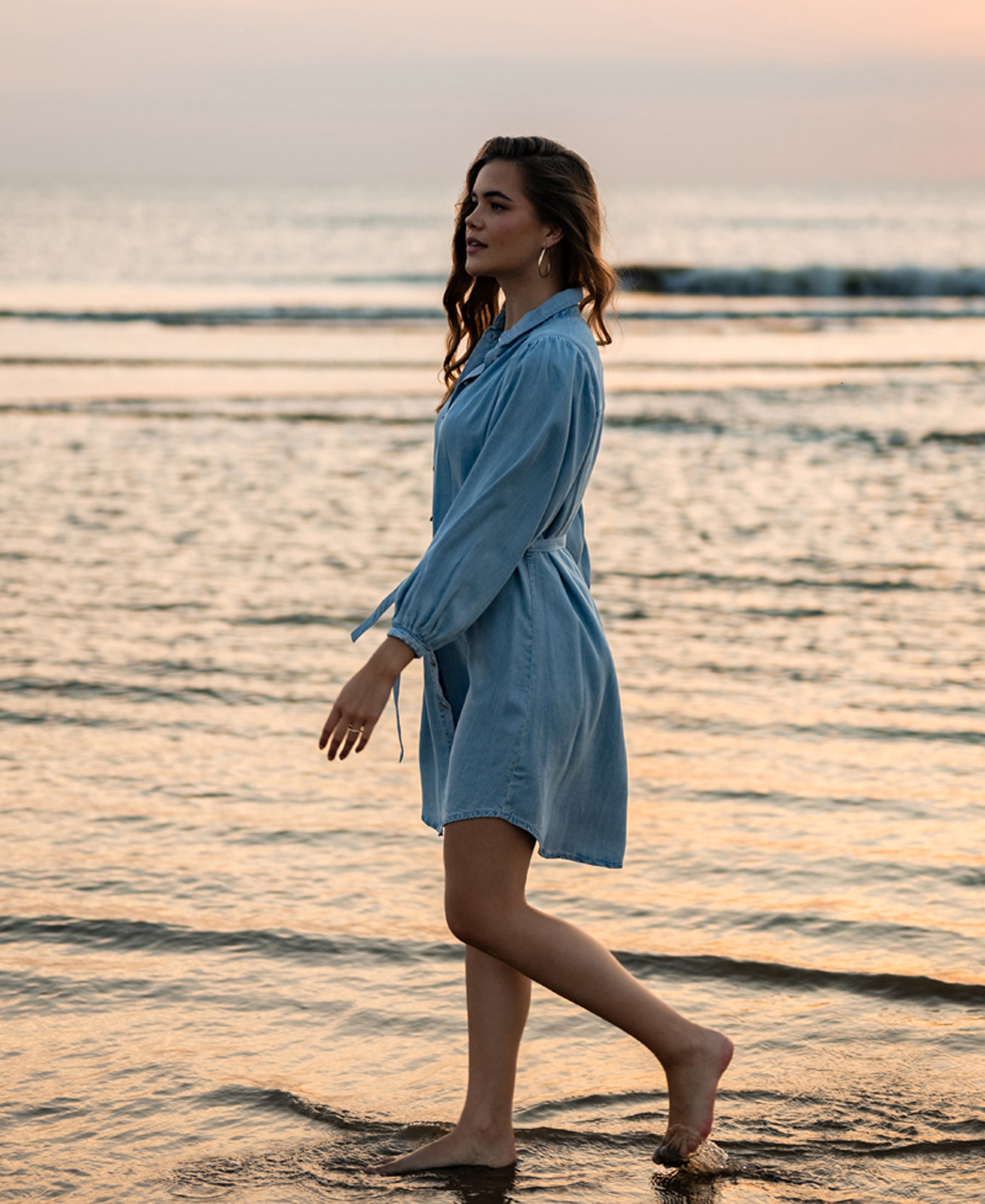 Woman wearing light blue Tencel shirt dress walks along shoreline at sunset, waves in the background.