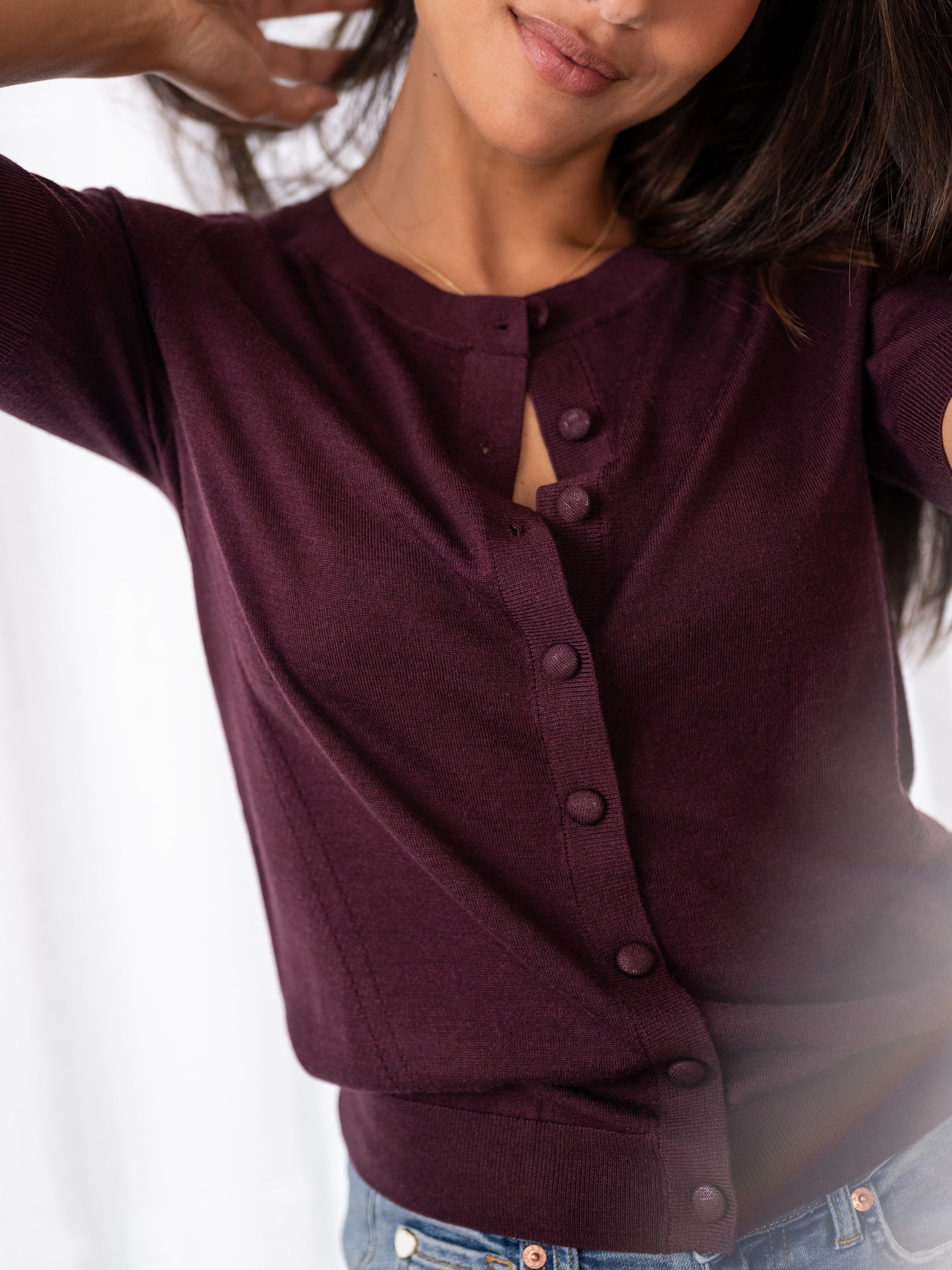 Woman in a PLEIN PUBLIQUE merino wool and silk cardigan, style LA LORAINE, color wine, arm raised against white background.