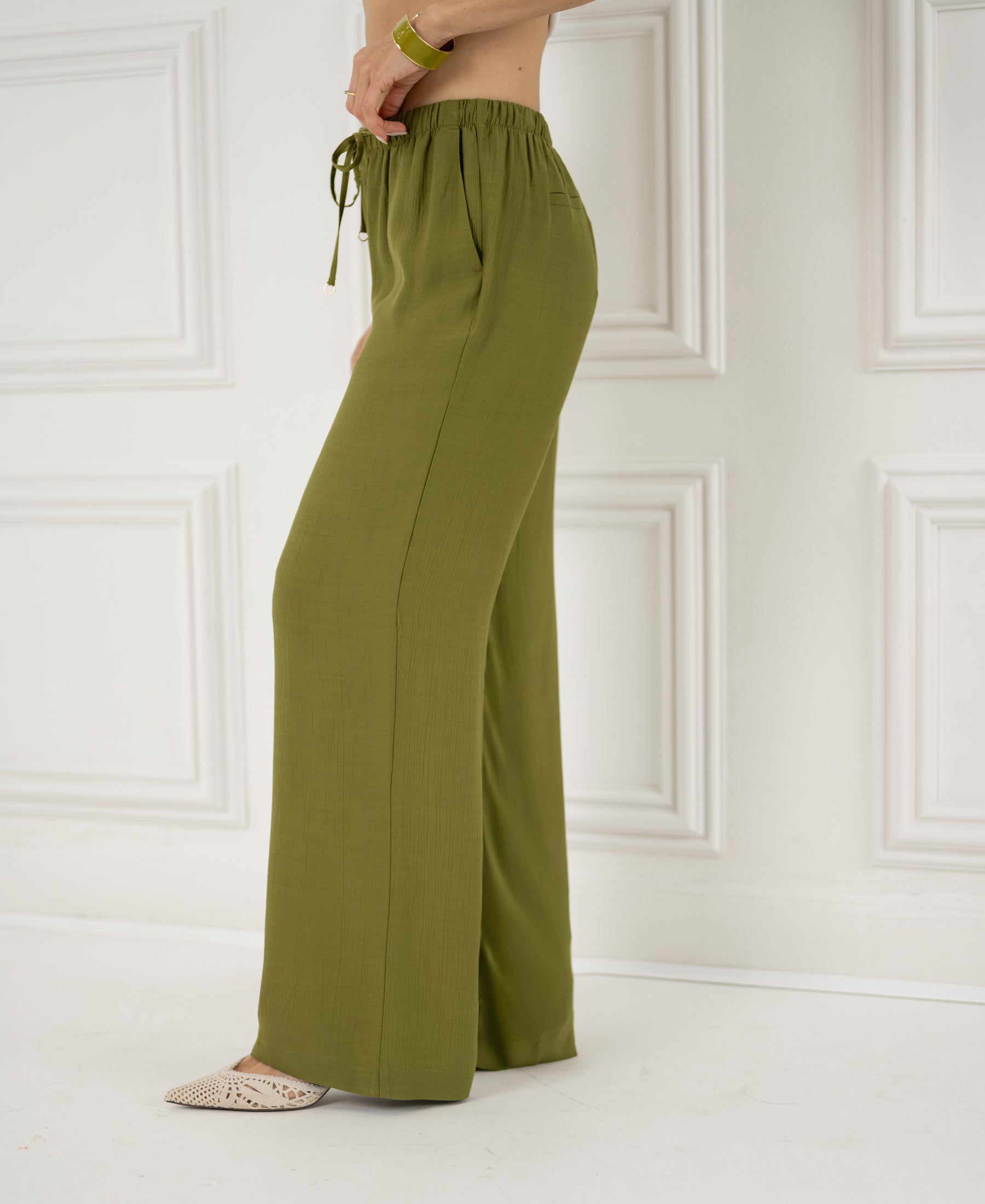 Olive green LA LYNETTE trousers shown against a white paneled wall.