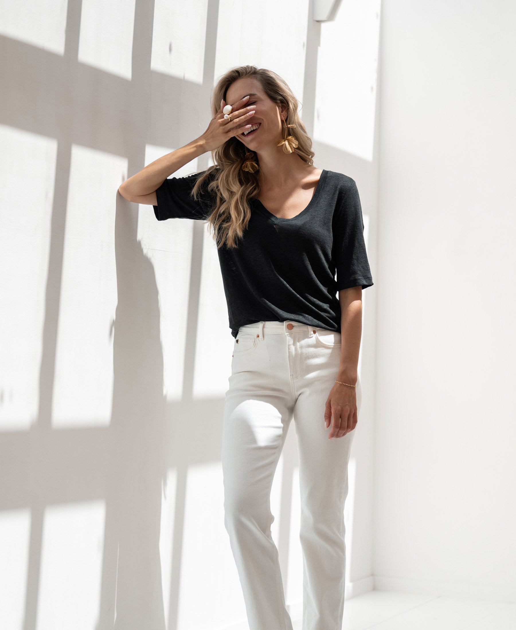 Woman wearing black linen T-shirt smiles in sunlight, hand covering her mouth.