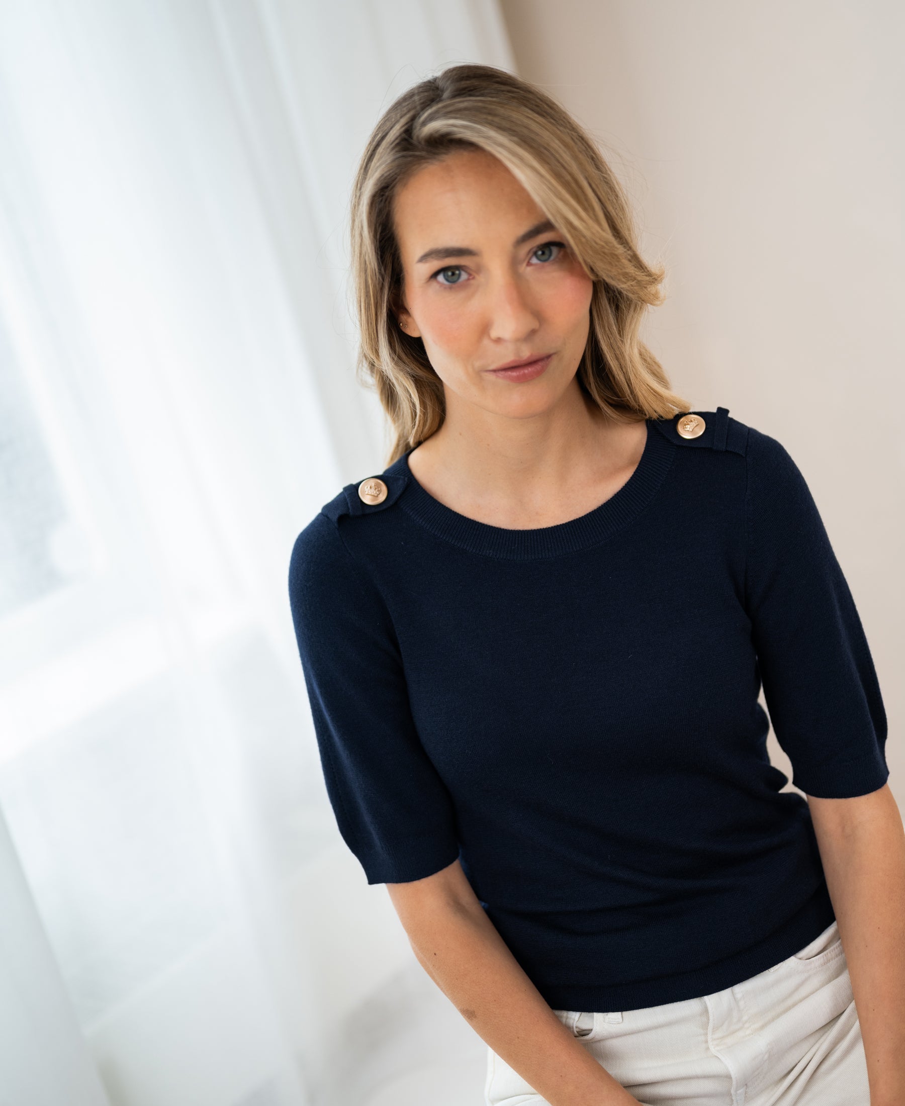 Navy blue merino wool sweater by PLEIN PUBLIQUE, shown on a model standing near a window with white curtains.