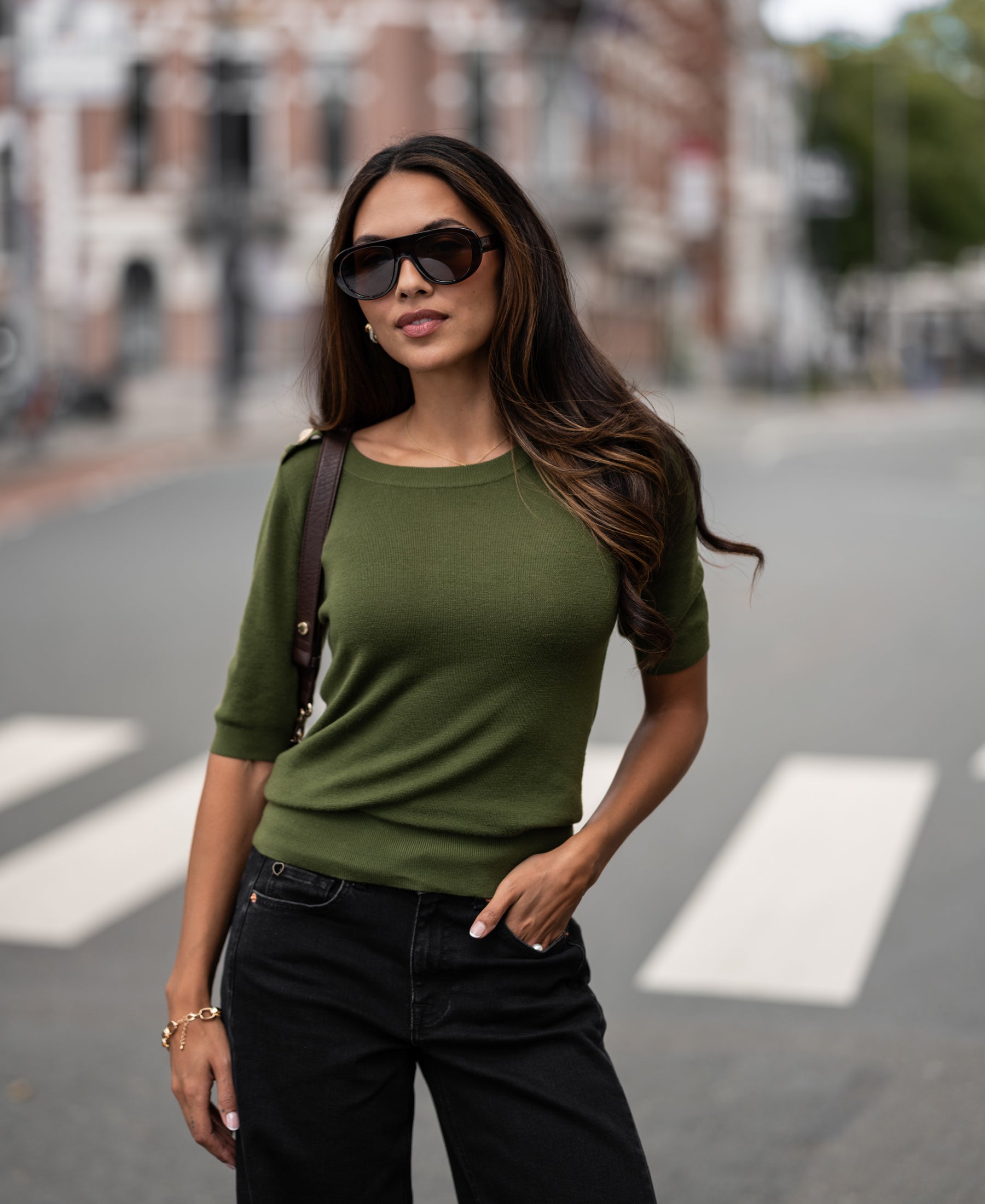 Woman wearing olive green LE MAJOR Merino sweater, hand in pocket, standing on a city crosswalk.