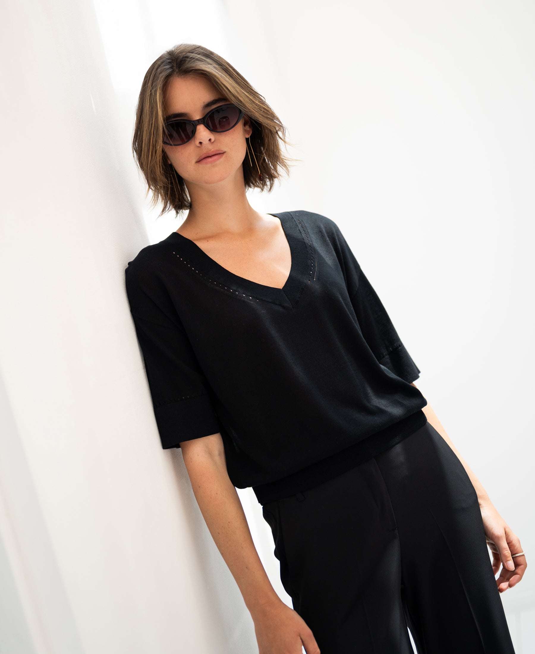 Black LE MARDY silk top by PLEIN PUBLIQUE displayed on model leaning against a white wall.
