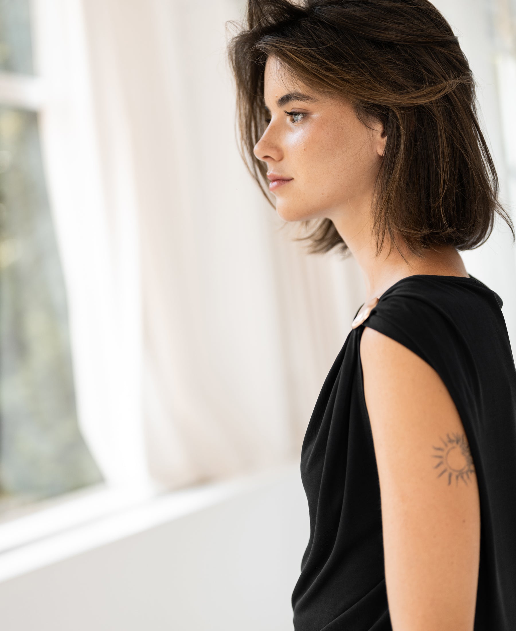 Woman wearing black LA MAYA top with buckle by PLEIN PUBLIQUE, sun tattoo visible on her shoulder.