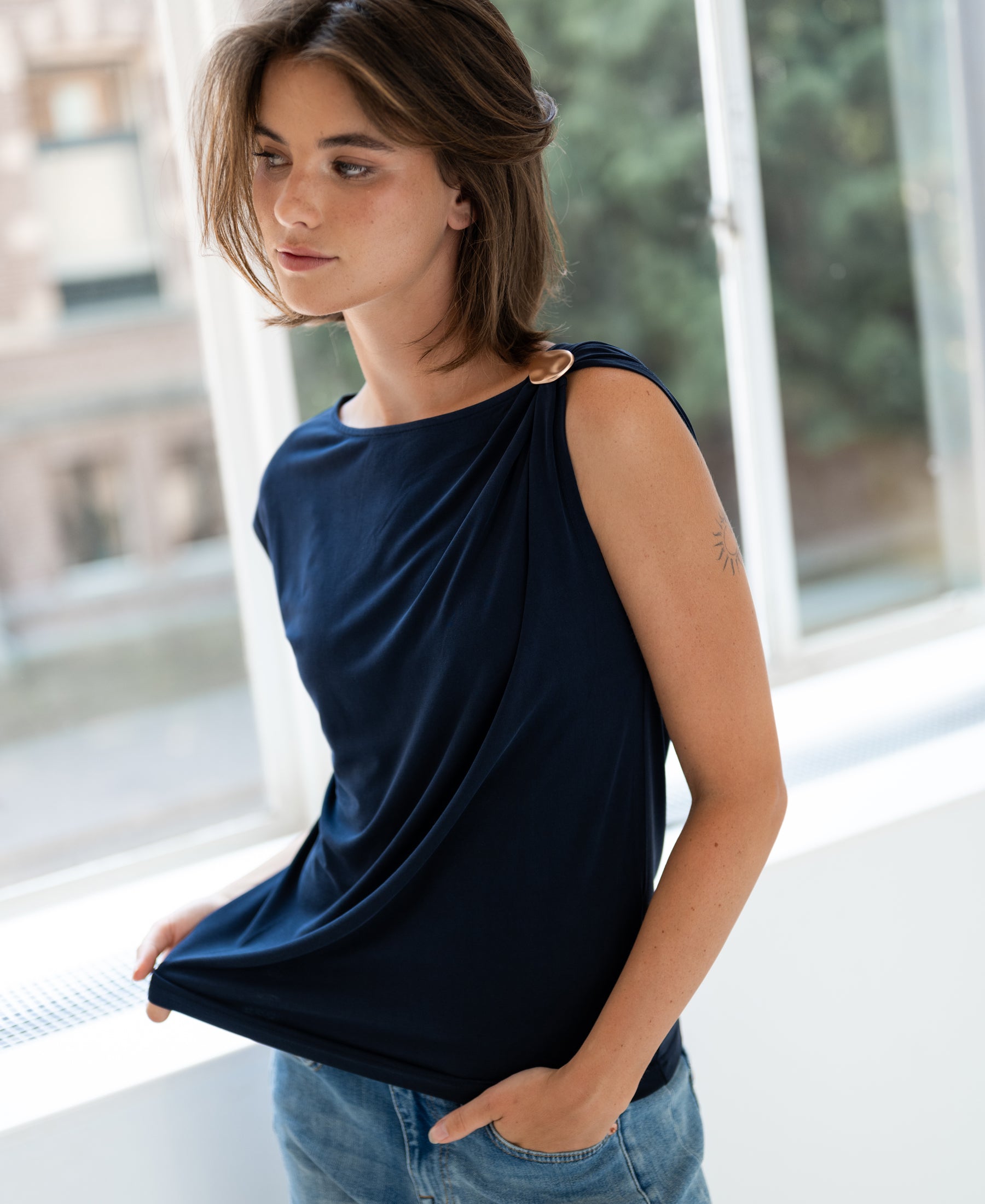 Woman wearing a PLEIN PUBLIQUE navy LA MAYA top with buckle, standing by a window and looking aside.