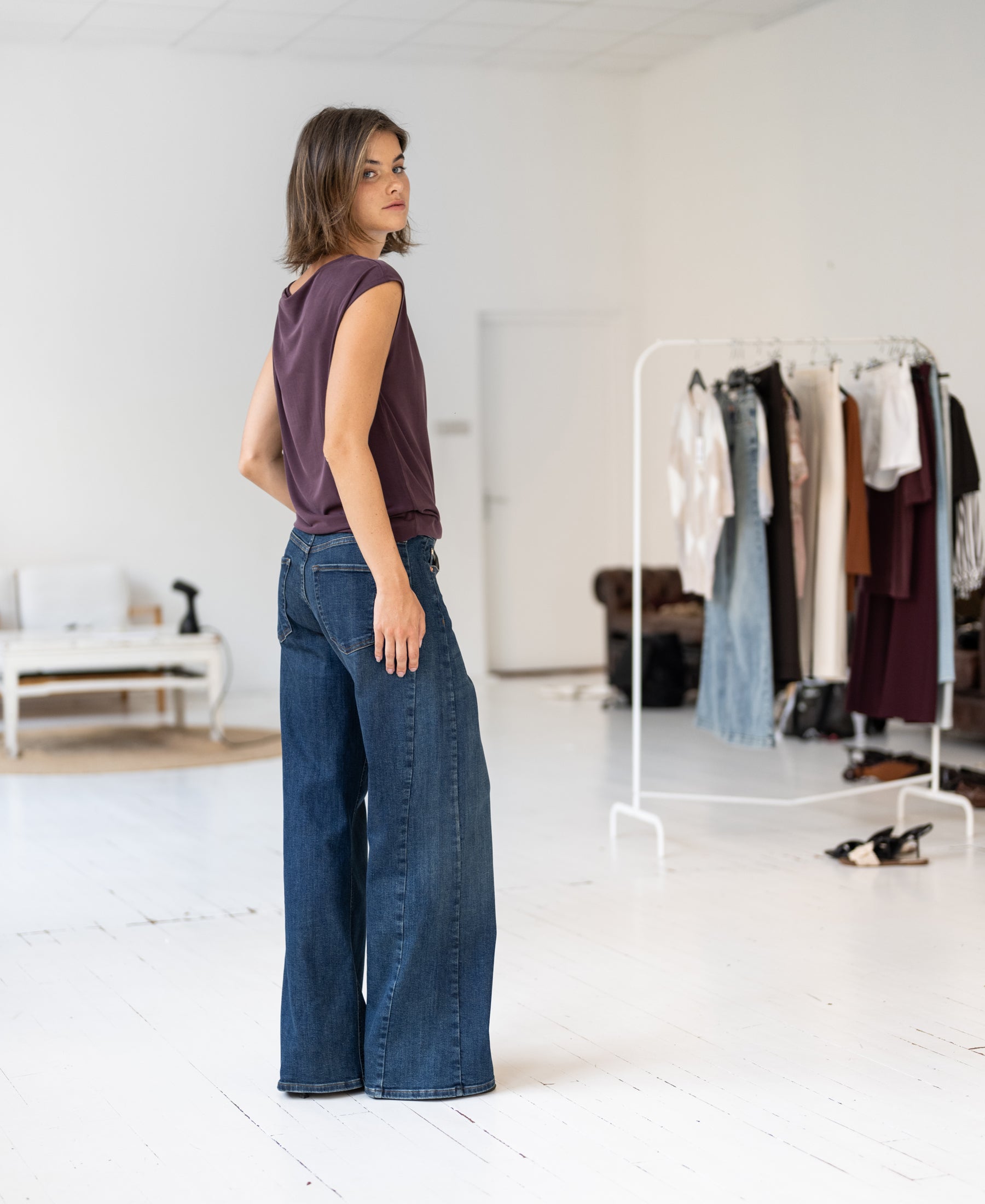 Woman wears the PLEIN PUBLIQUE LA MAYA Wine Belted Top, standing by a rack of clothes.