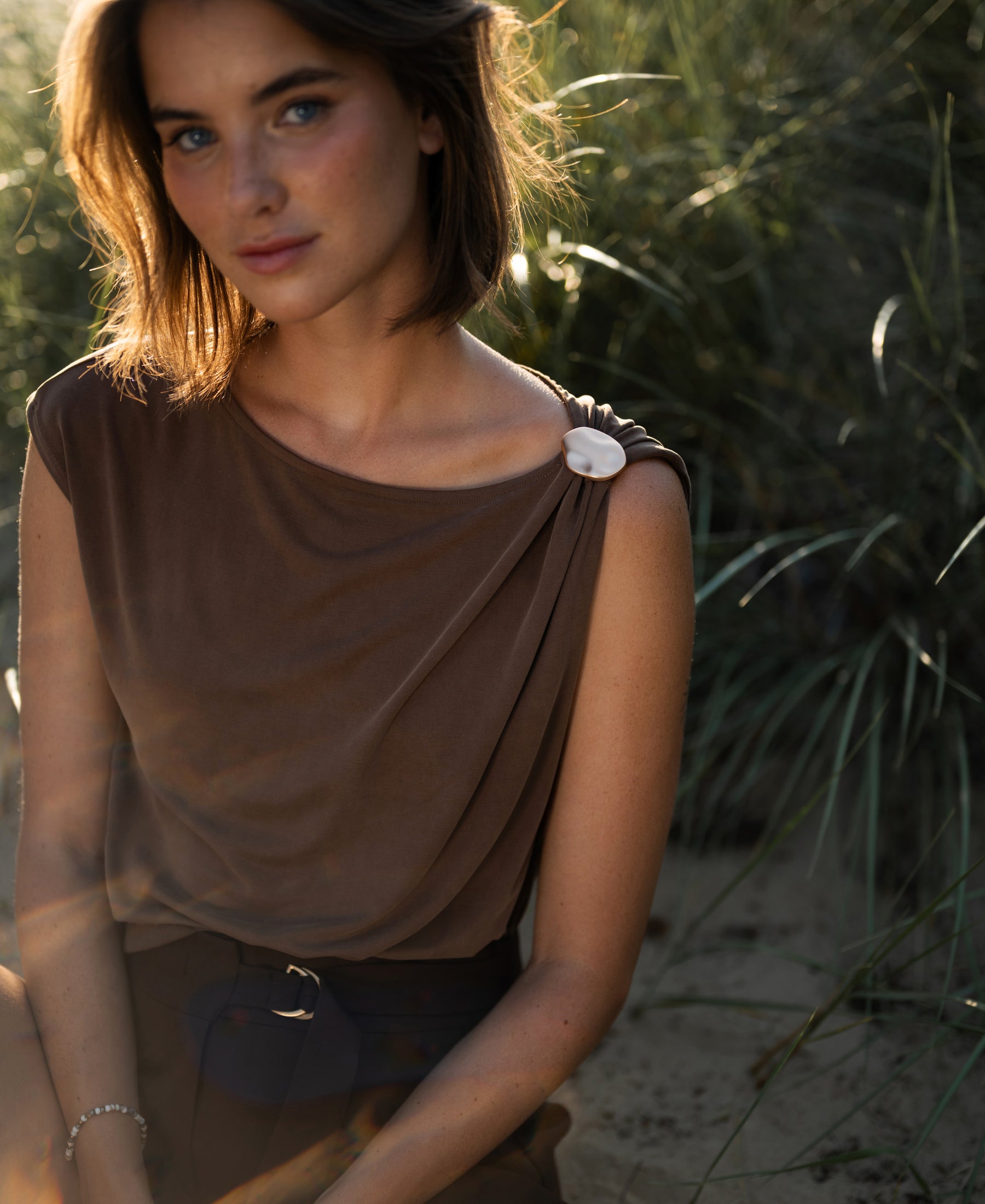 Brown LA MAYA top with buckle by PLEIN PUBLIQUE, shown outdoors among sunlit tall grass.