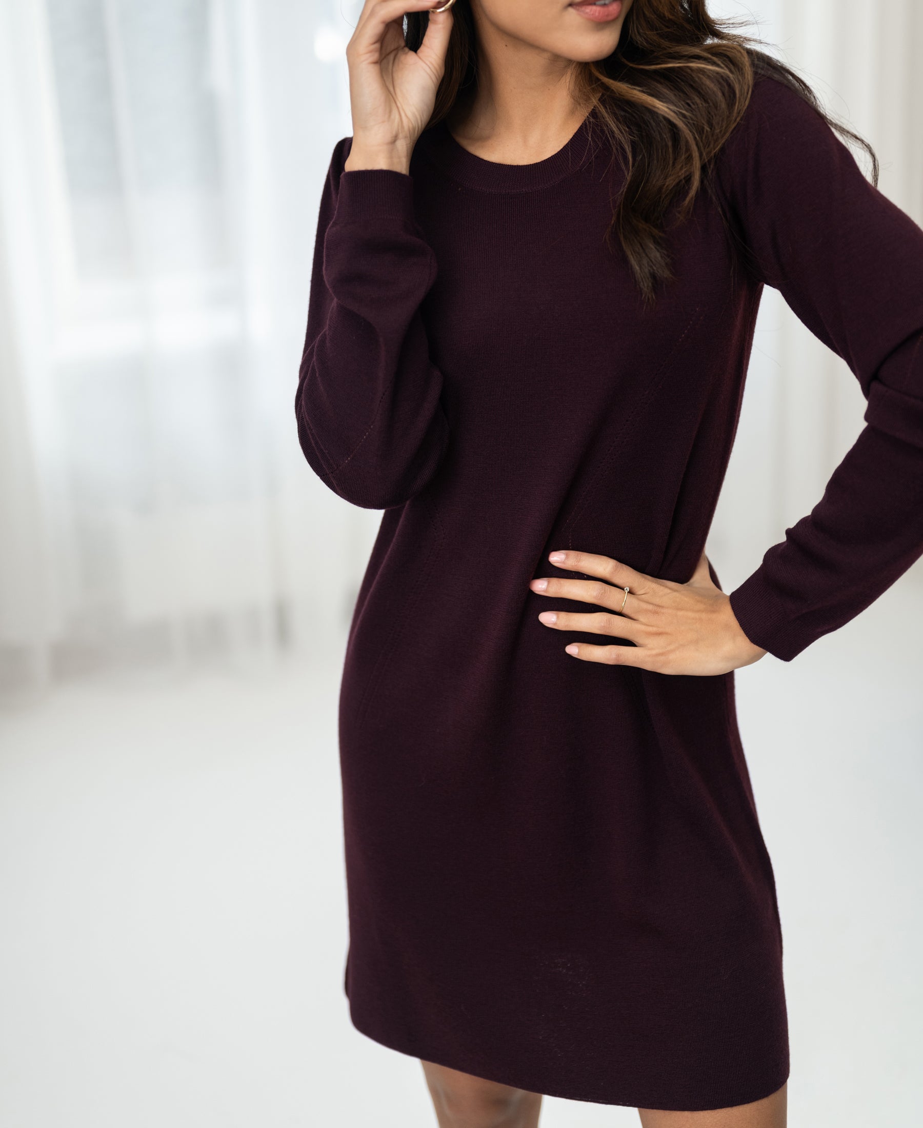 Woman indoors wearing the LA MIMI Merino wool dress in aubergine from PLEIN PUBLIQUE, hand near ear and hip.