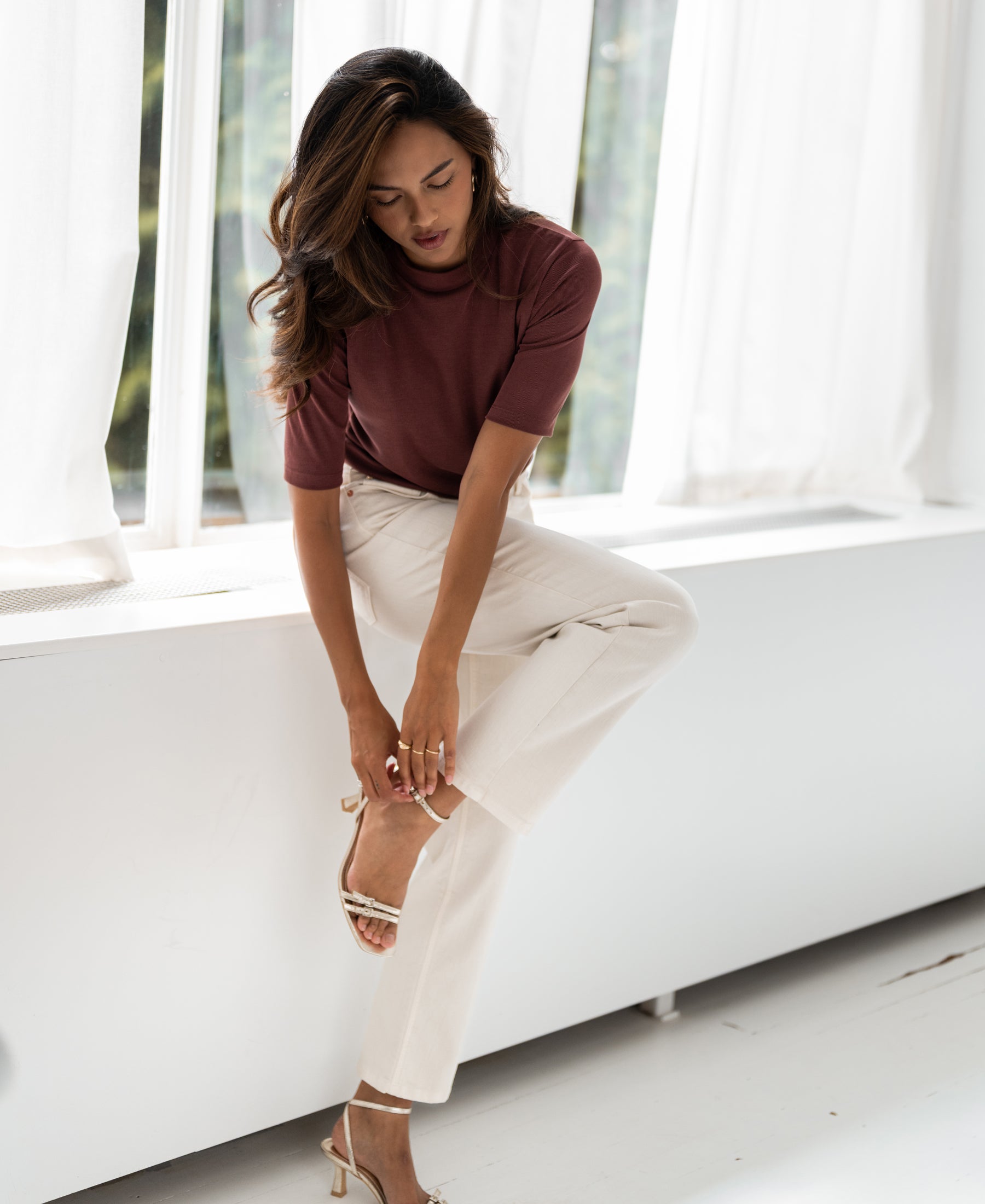 Woman wearing a ribbed cacao-colored top from PLEIN PUBLIQUE, sitting on a windowsill.