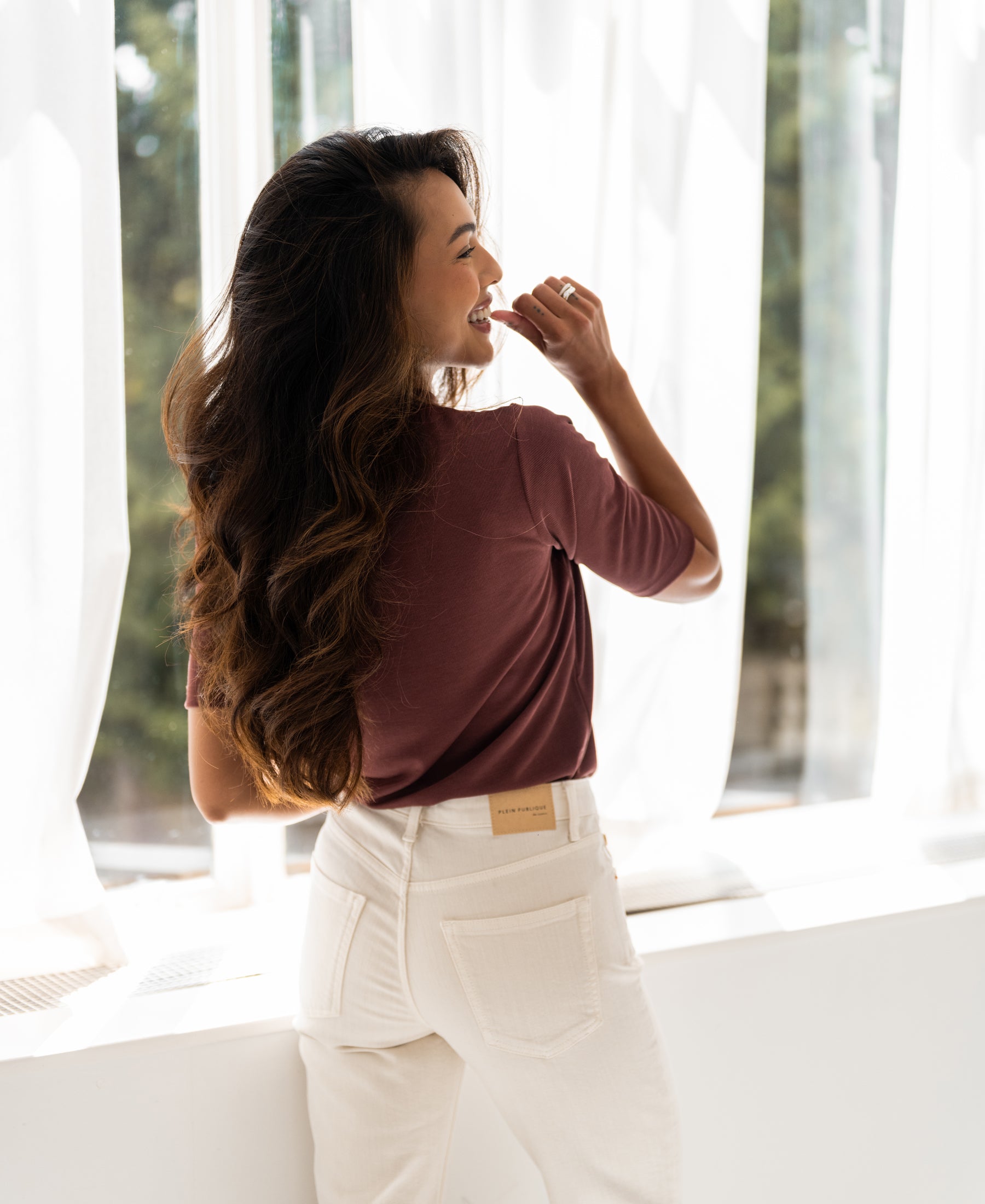 Woman wearing a ribbed cacao-colored top stands by a sunlit window, smiling and looking outside.