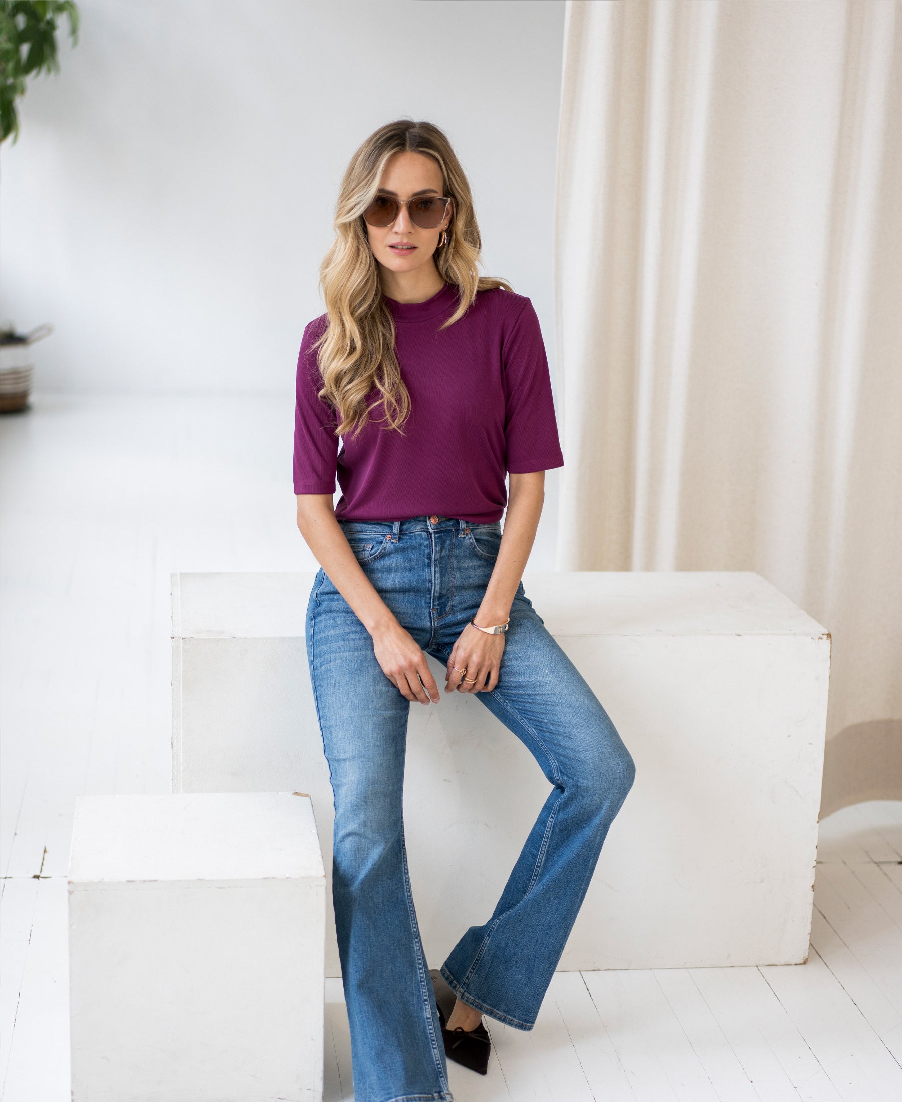 Woman wearing light blue flared jeans sits on a white block in a bright room.