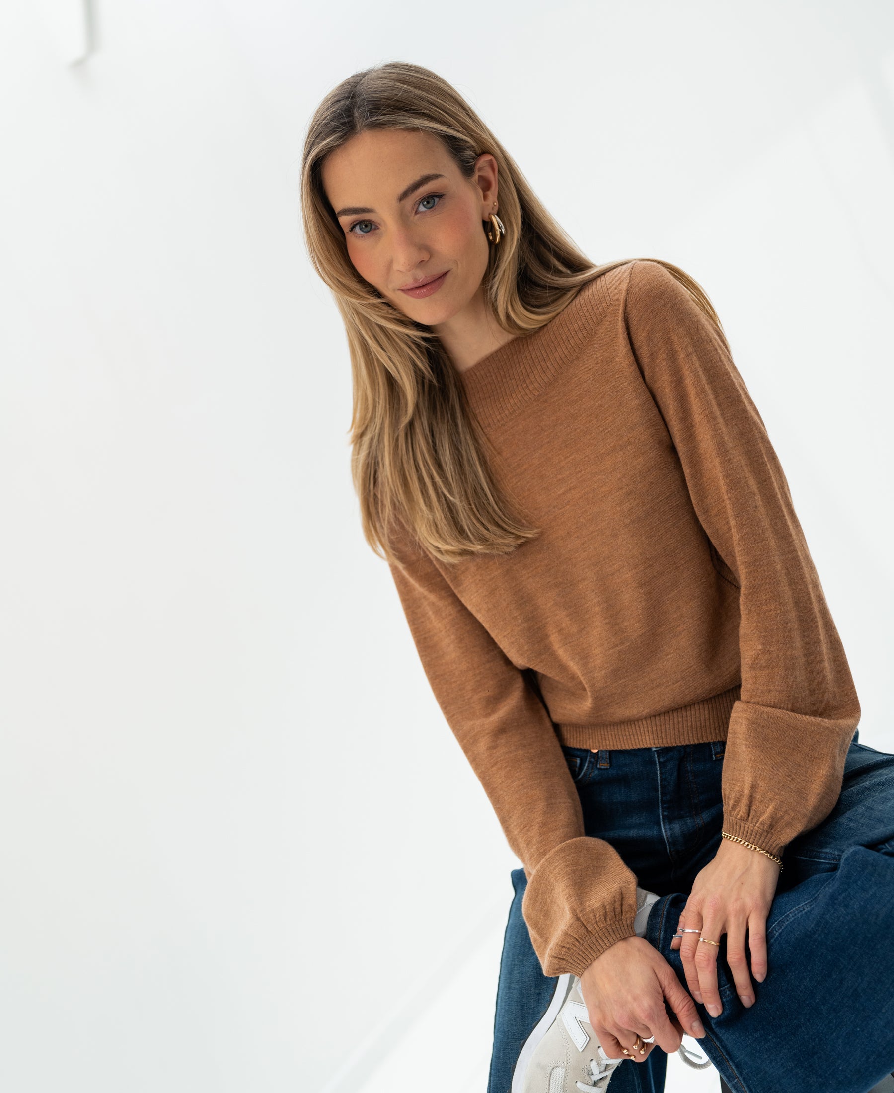Woman wearing PLEIN PUBLIQUE’s LA MUSE boat neck sweater in chai tea color, standing in a bright, minimalist space.
