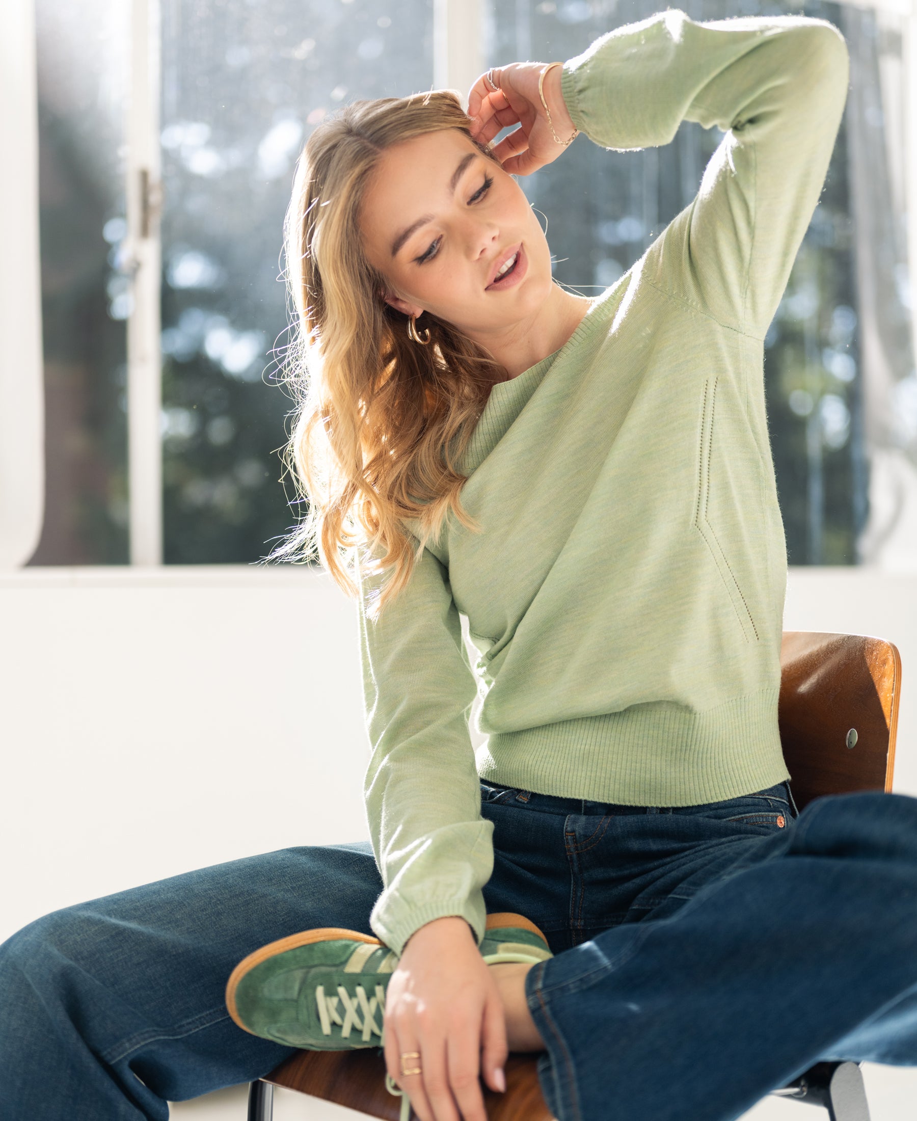 Woman wears the PLEIN PUBLIQUE boat neck sweater in light green, sitting on a wooden chair in sunlight.