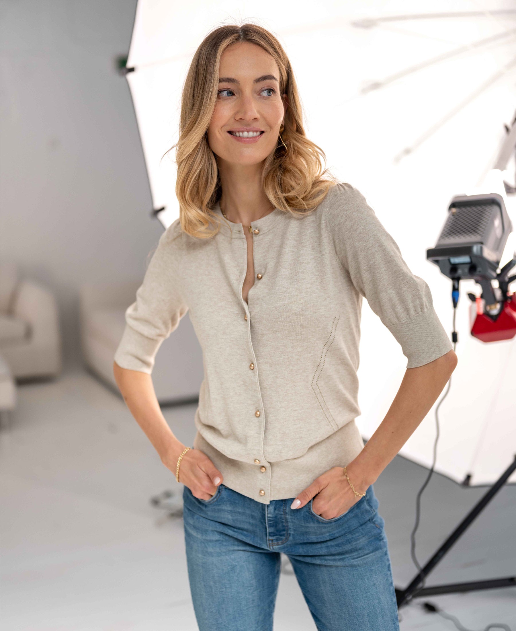 Woman wearing the PLEIN PUBLIQUE ADELINE cardigan in sand-gold, standing in a photo studio with lights behind her.