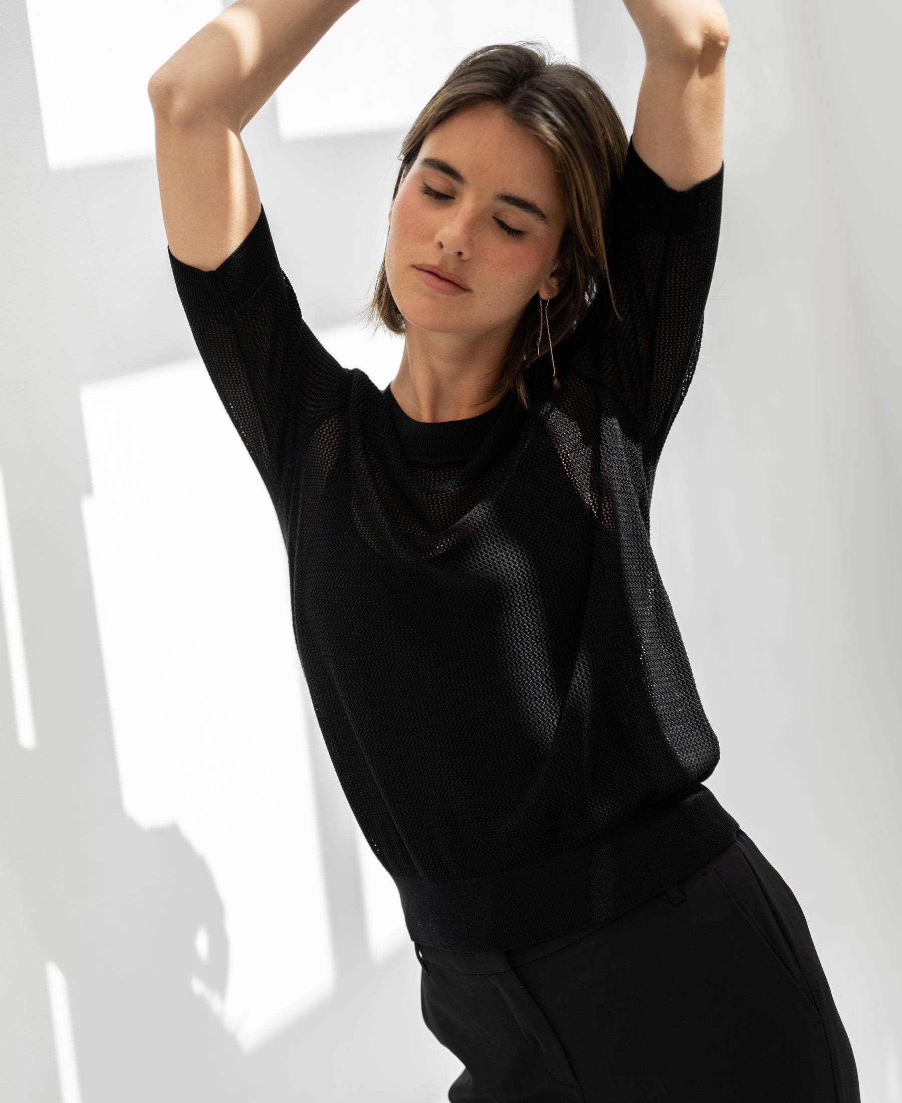 Black mesh sweater with silk by PLEIN PUBLIQUE shown on a woman, eyes closed, sunlight on the wall behind.