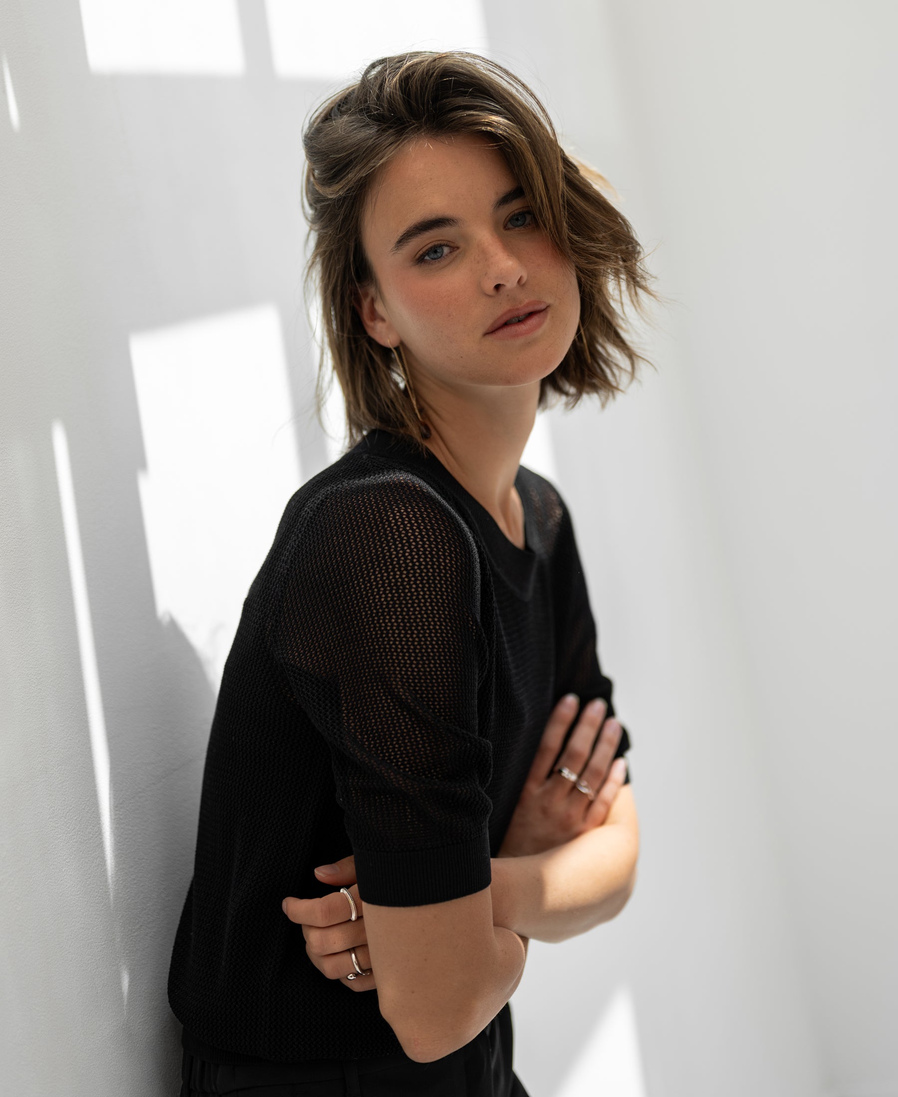 Black mesh sweater with silk, front view. Model stands by a sunlit white wall with arms crossed.