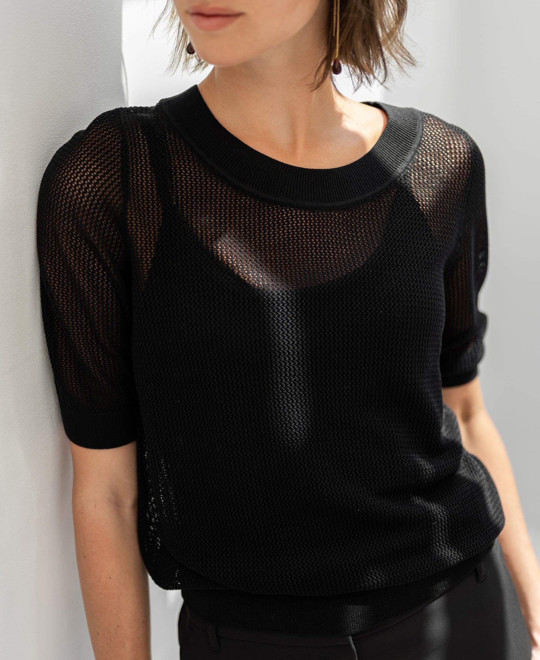 Woman wears black mesh silk sweater by Plein Publique, standing against a white wall.