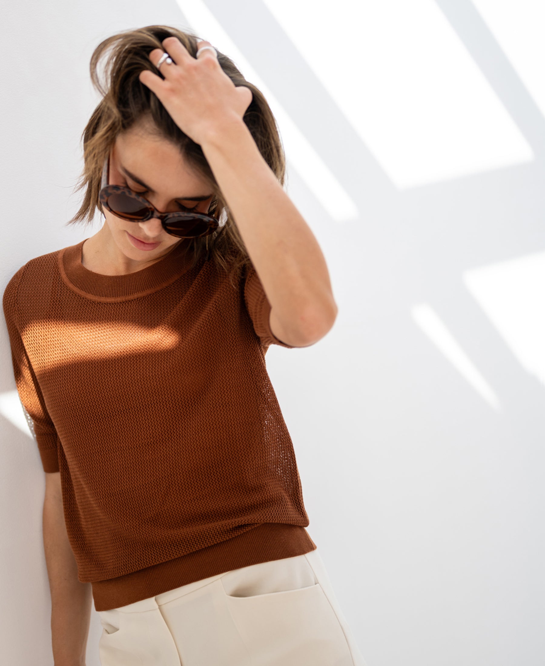 Woman wearing the PLEIN PUBLIQUE mesh sweater LE NANCY with silk in Havana color, standing against a white wall, brushing her hair.