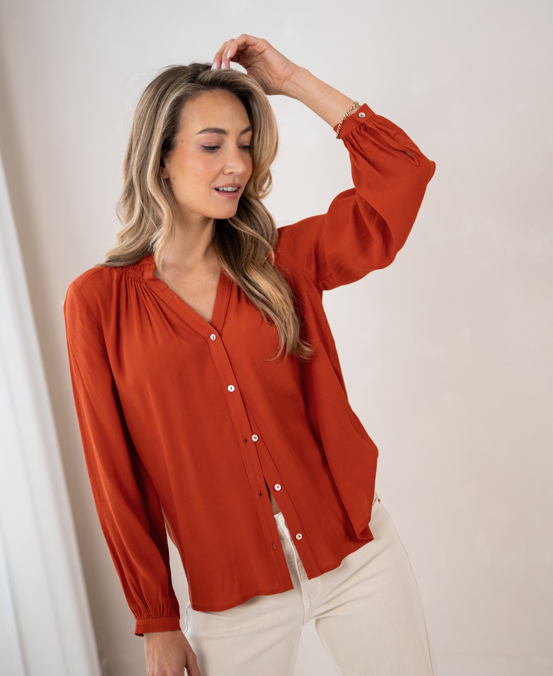 Woman wearing a burnt orange blouse, smiling softly and touching her head.