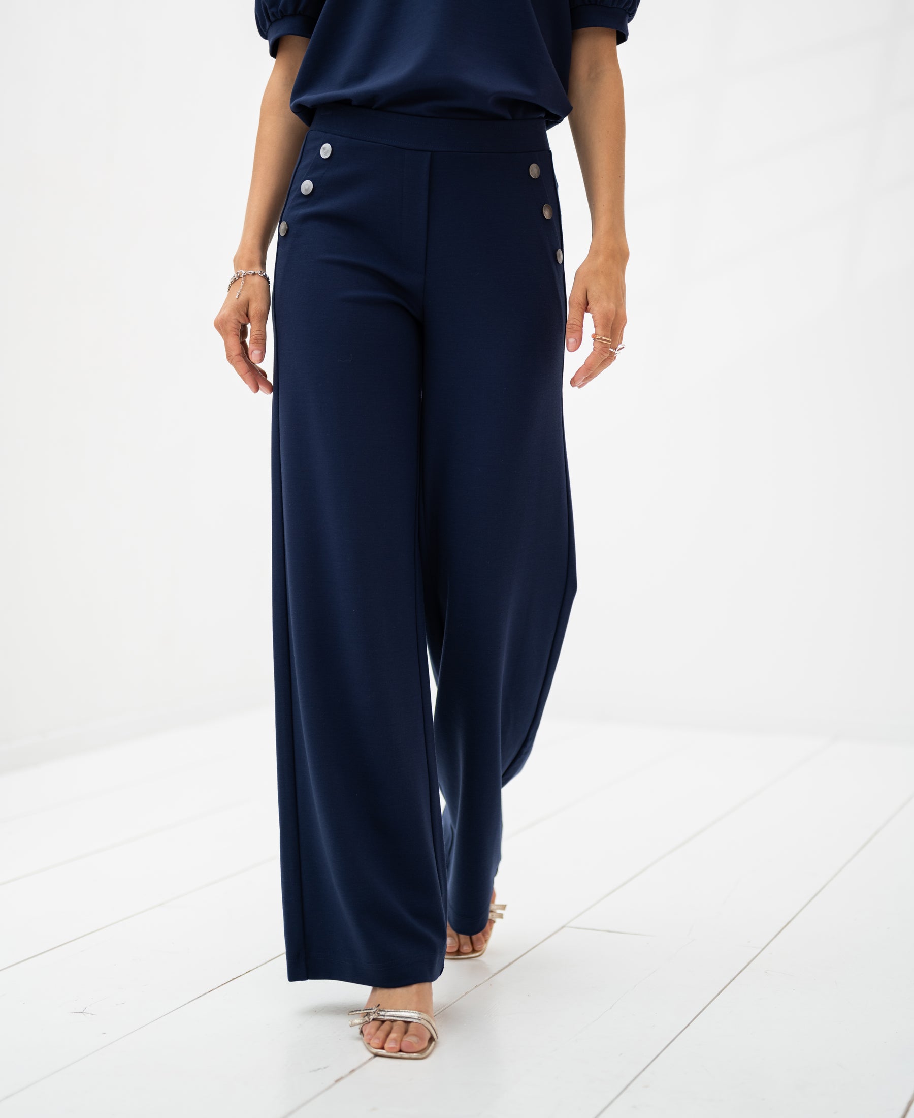 Dark blue deluxe travel pants, shown from shoulders down, displayed on a white floor.
