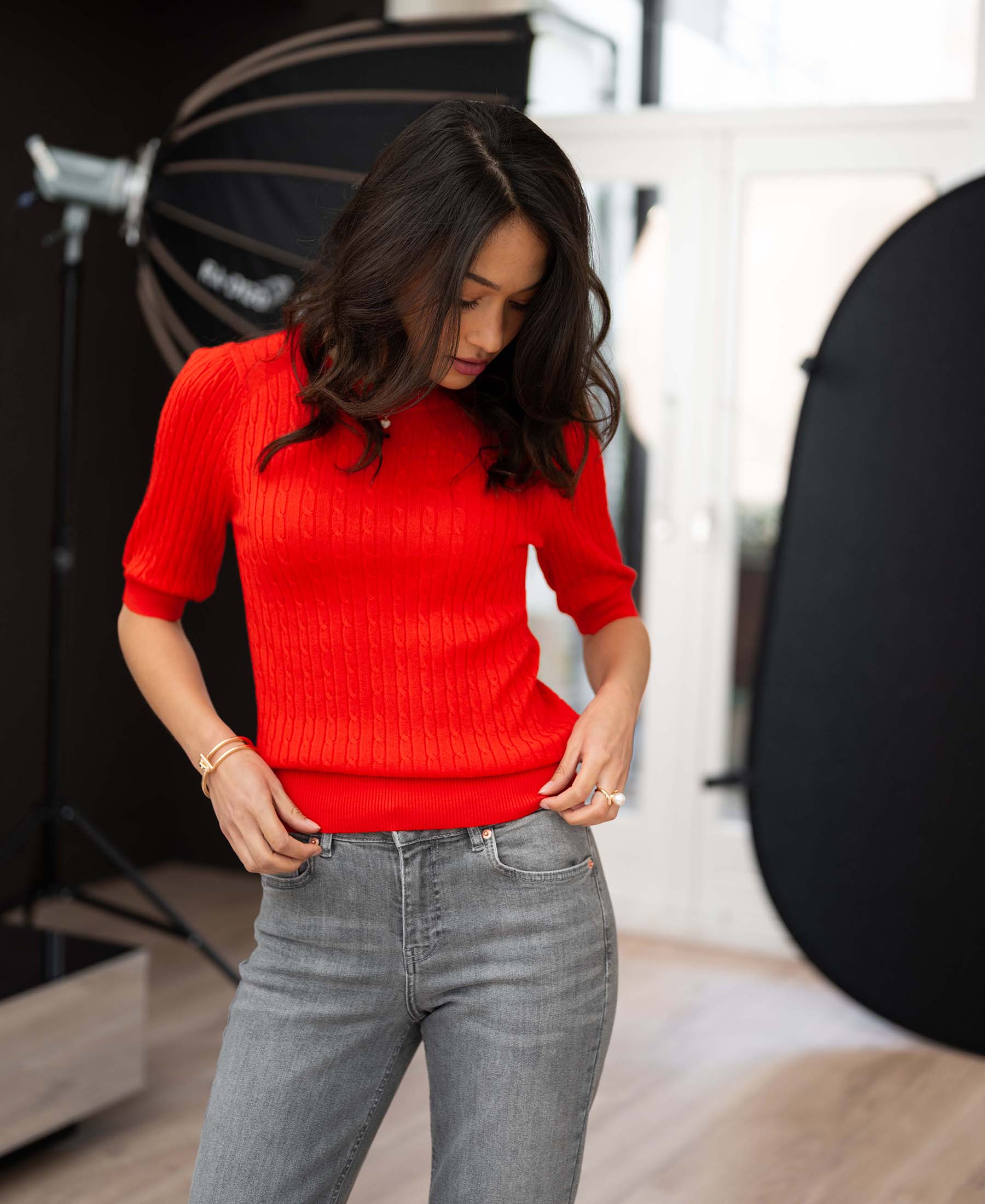 Woman wears coral-red cable knit short-sleeve sweater, adjusts it while standing in studio.