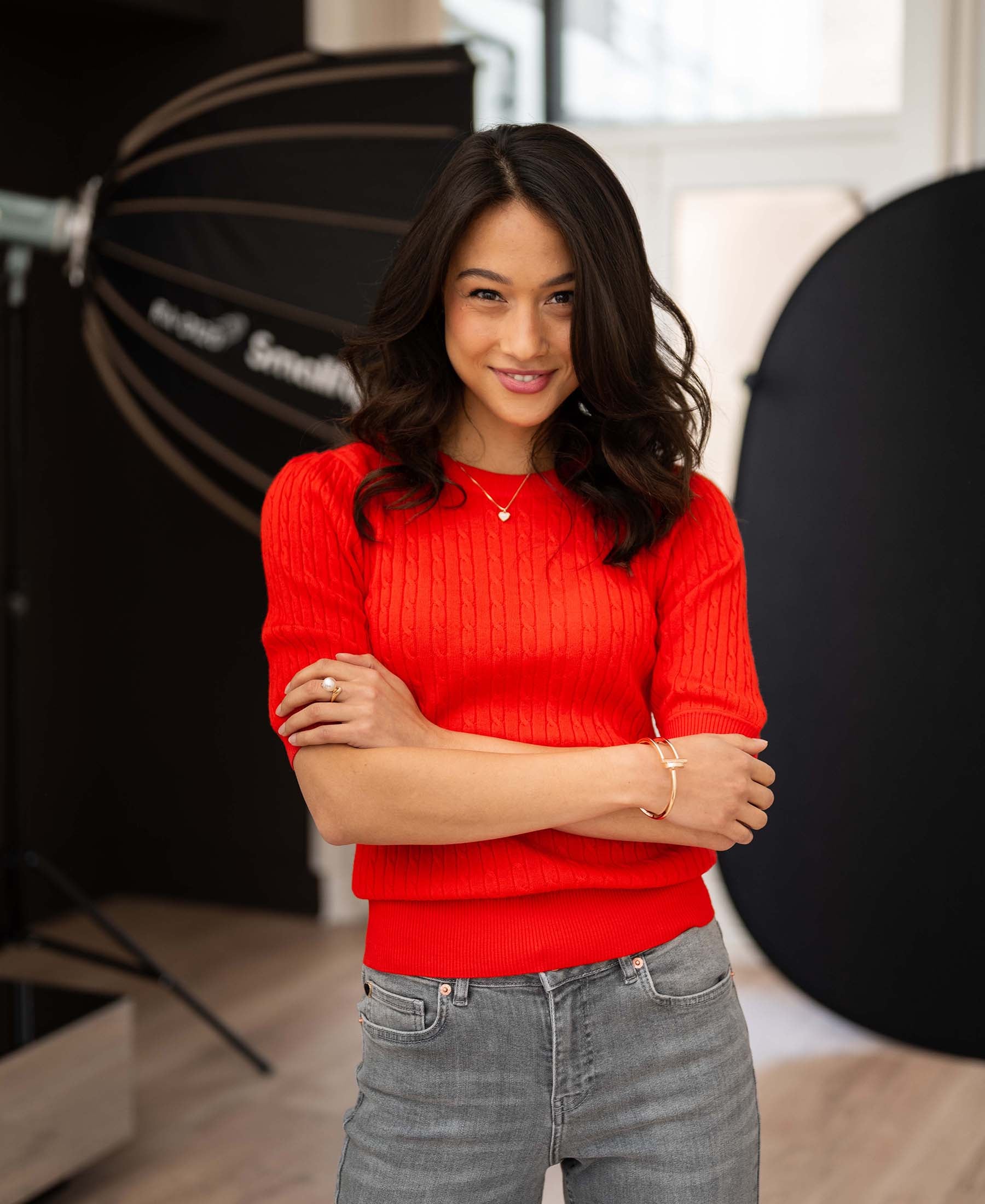 Coral red short-sleeve cable knit sweater by LA NONNA, shown on a model standing with arms crossed in a studio.