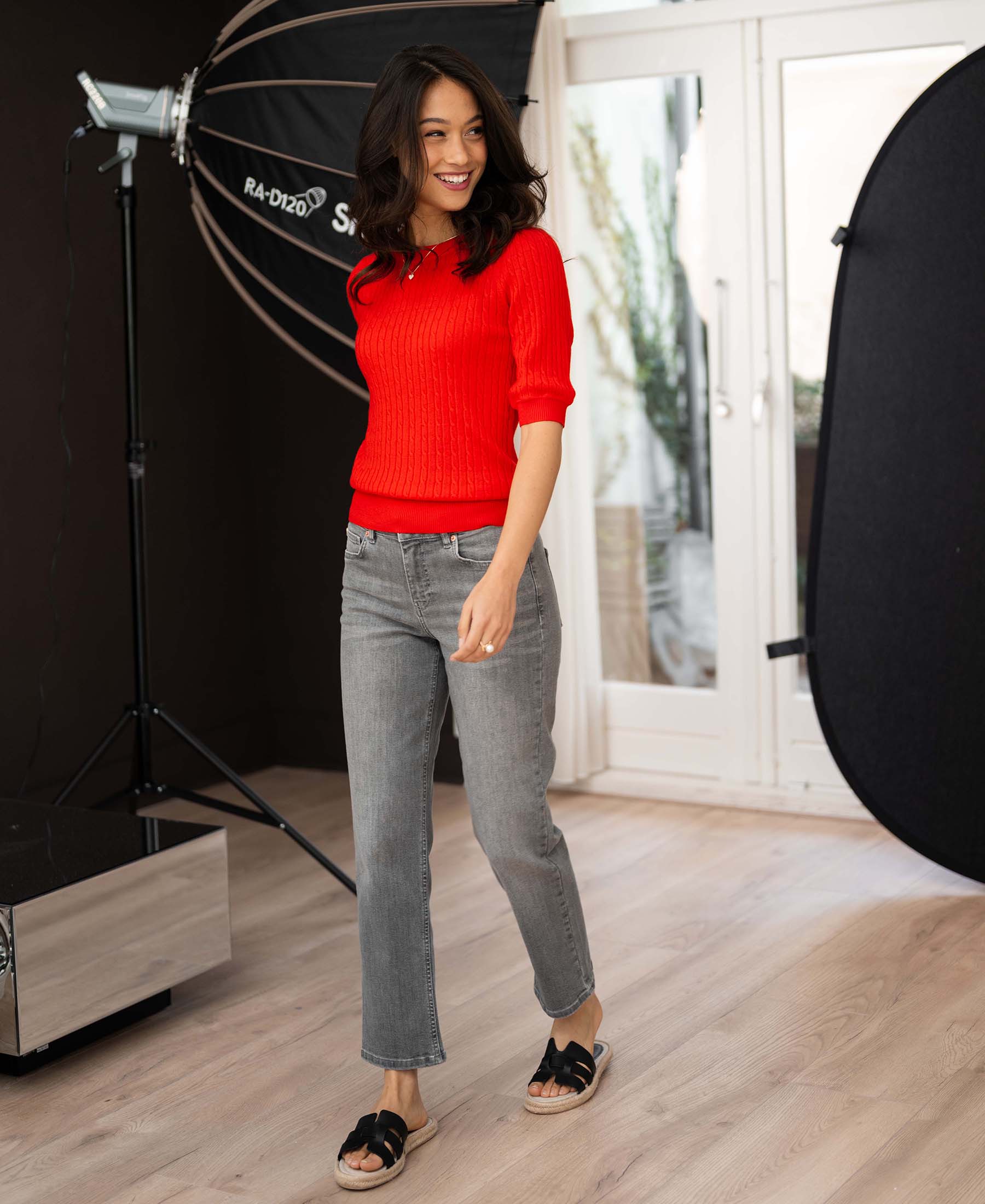 Woman wearing a coral red short sleeve cable knit sweater, walking in studio.