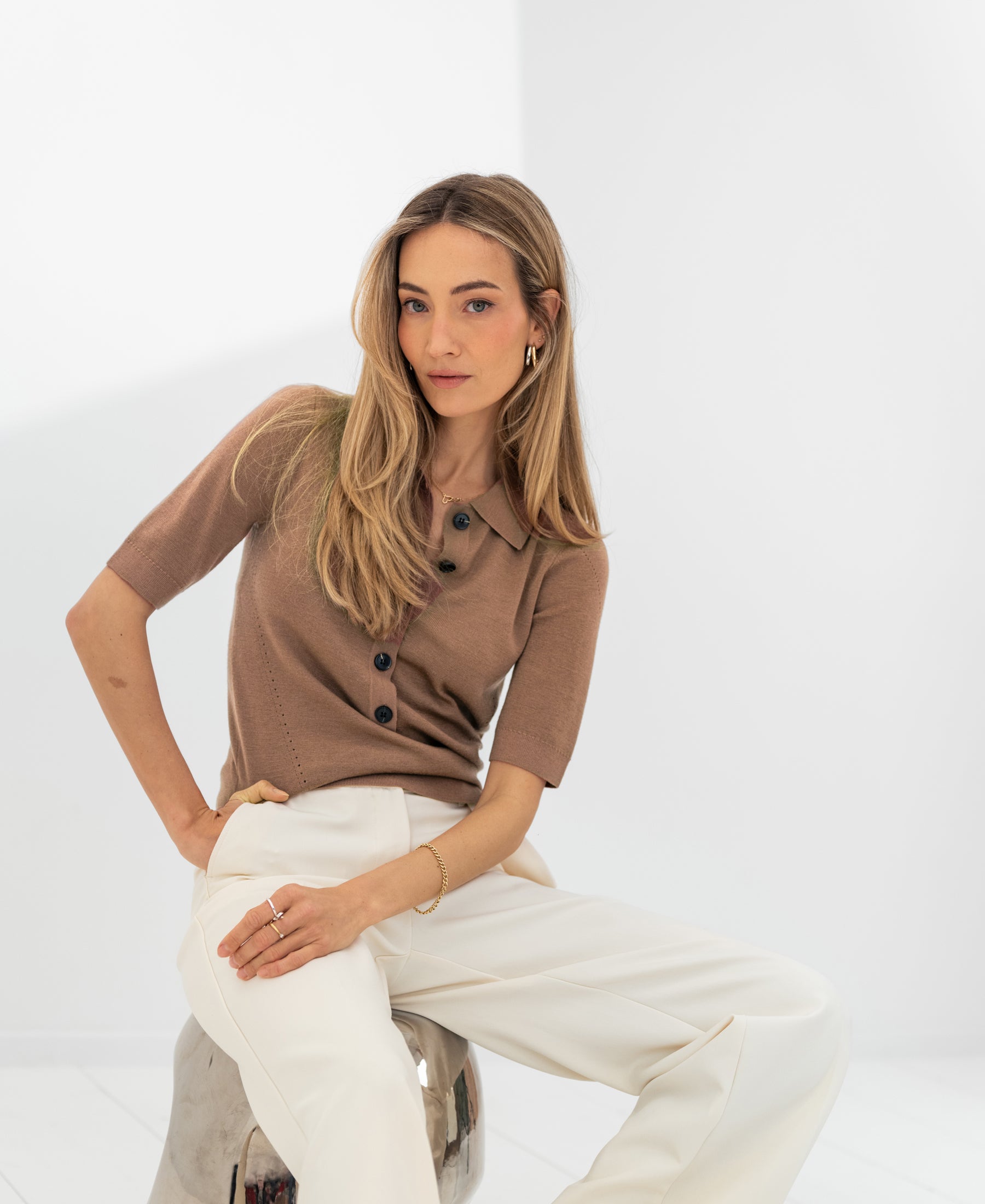 Soft camel-colored merino wool polo shirt by PLEIN PUBLIQUE, shown worn by a woman seated on a stool.